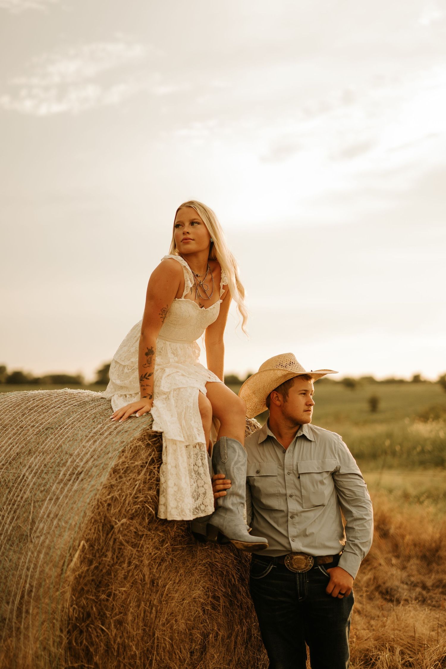 A woman in a white dress sits on a hay bale beside a man in a cowboy hat in a golden field.