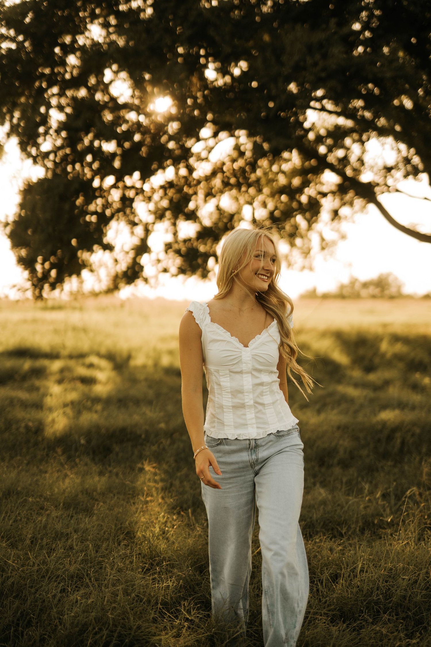 Blonde woman in white lace top and jeans smiling outdoors at golden hour with sunlight filtering through trees.