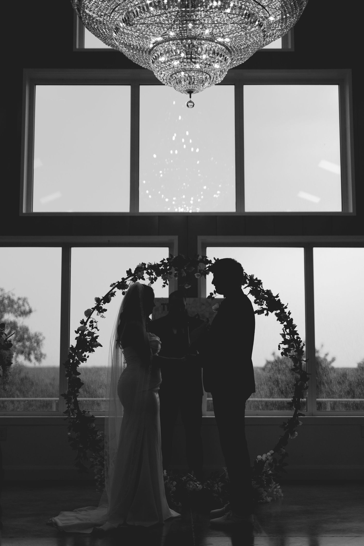 Black and white silhouette of bride and groom standing under floral arch with crystal chandelier above.