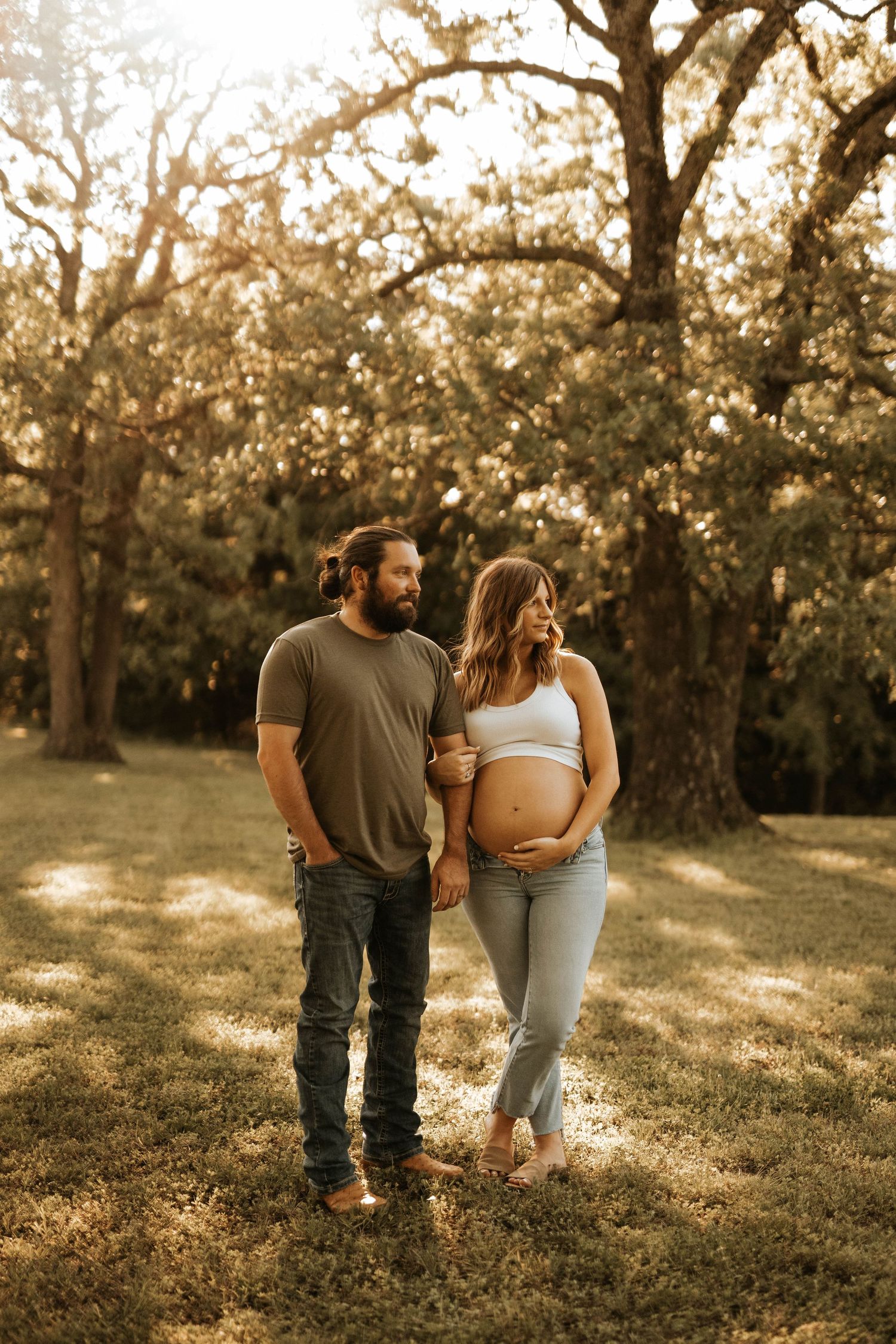 Couple walking through a sun-dappled park during golden hour maternity photoshoot.