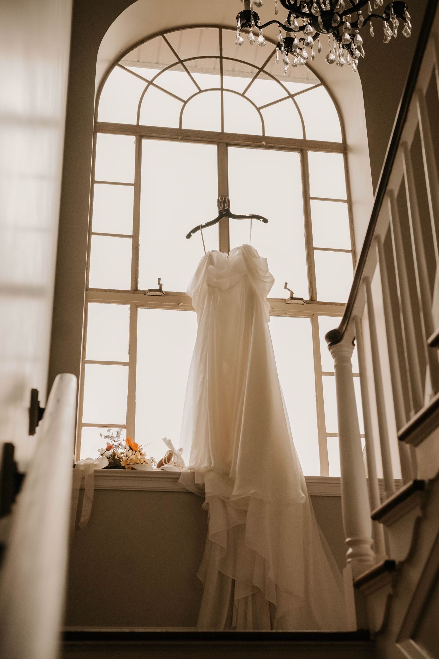 A white wedding dress hangs elegantly in front of an arched window with natural light.