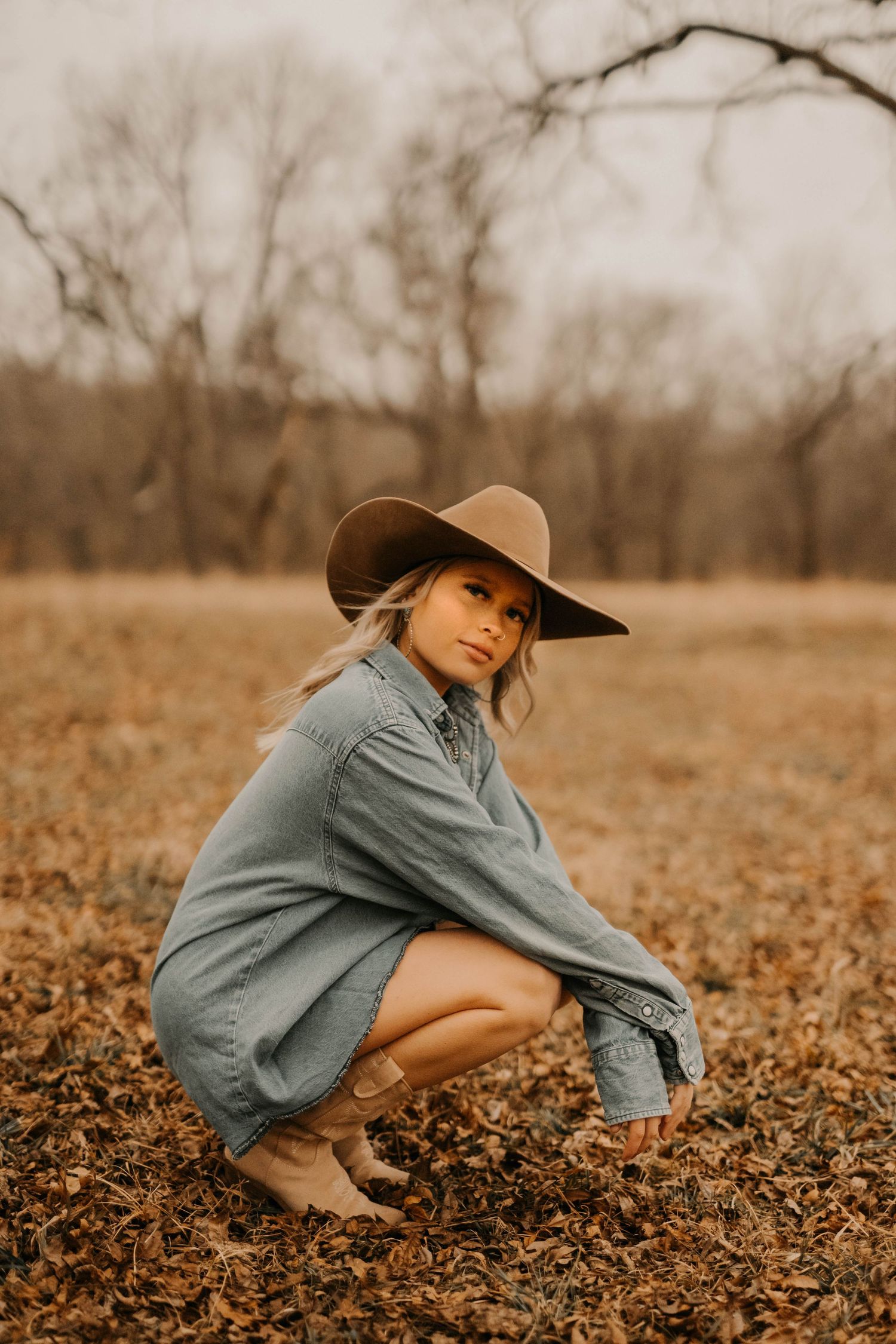 Woman in denim dress and wide-brim hat crouches in autumn field.