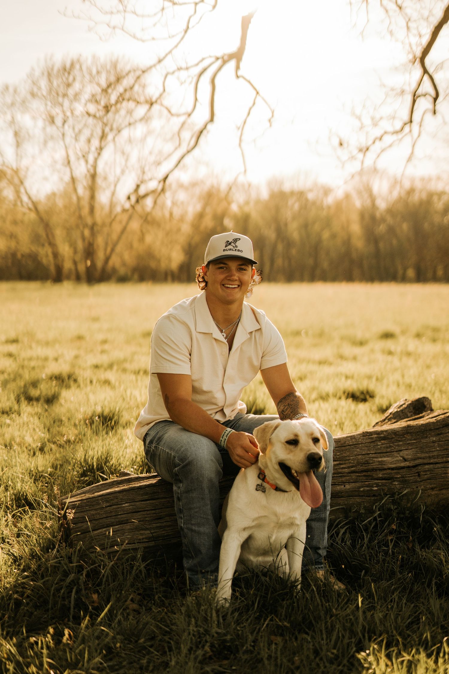 Young man in white shirt and cap sits on a log with a yellow Labrador in a sunny green field.