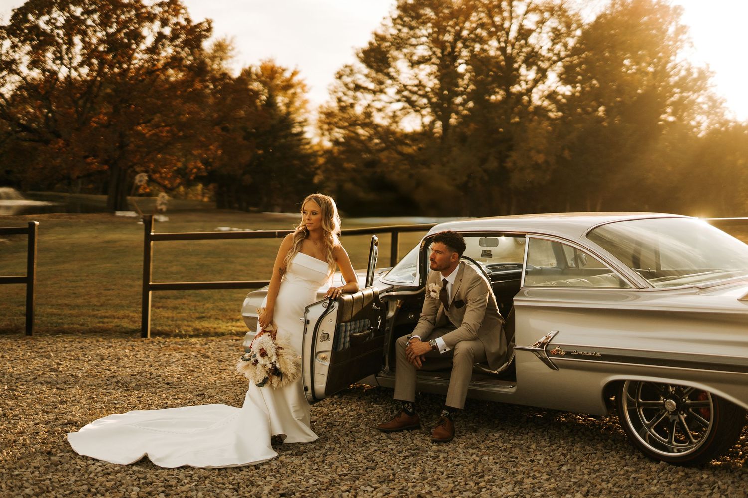 Bride in white gown and groom in gray suit pose beside a vintage silver car at golden hour outdoors.