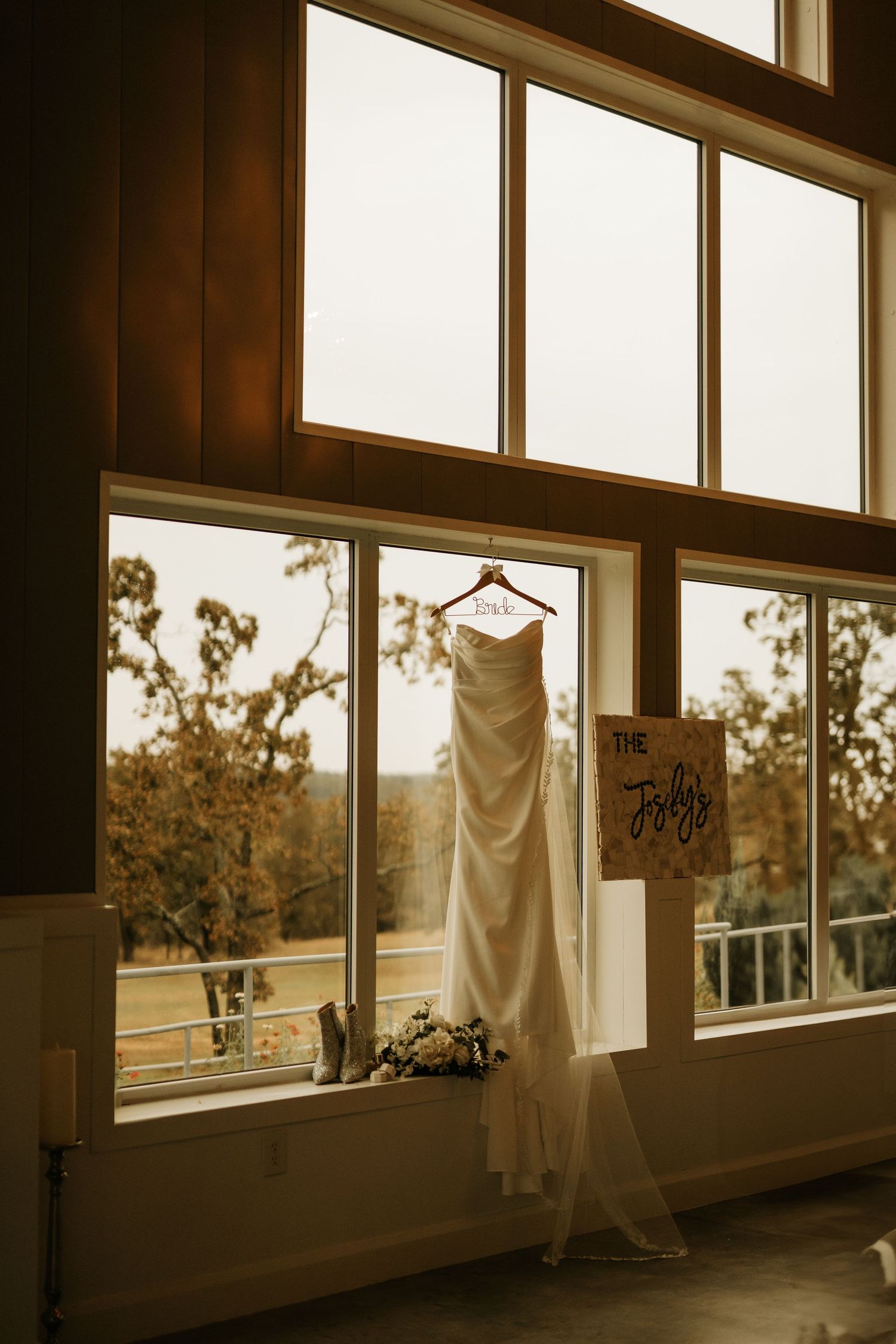 White wedding dress hanging on a wooden hanger in front of large windows with autumn trees visible outside.
