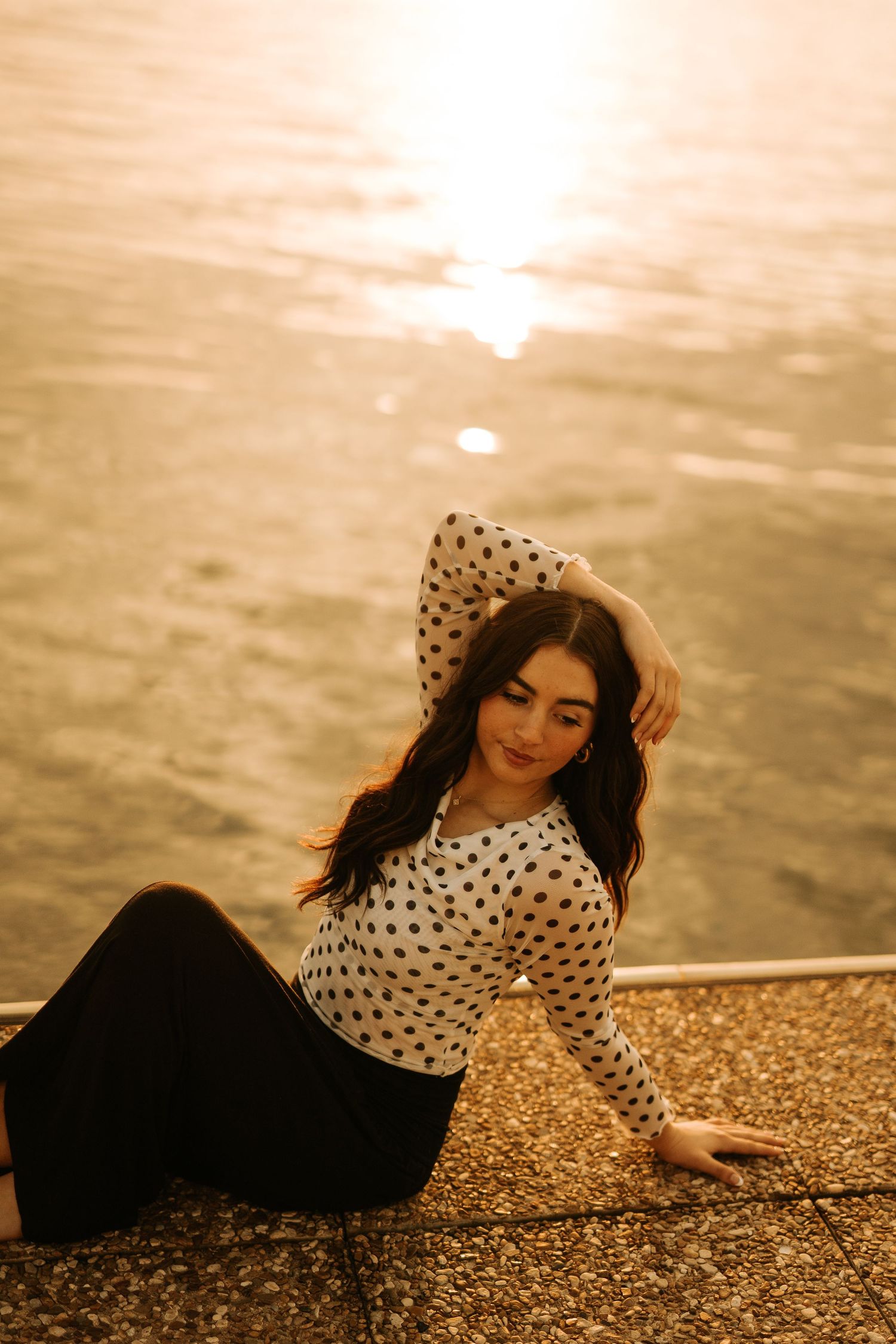 Young woman in polka dot top and black pants poses gracefully by shimmering golden sunset water.