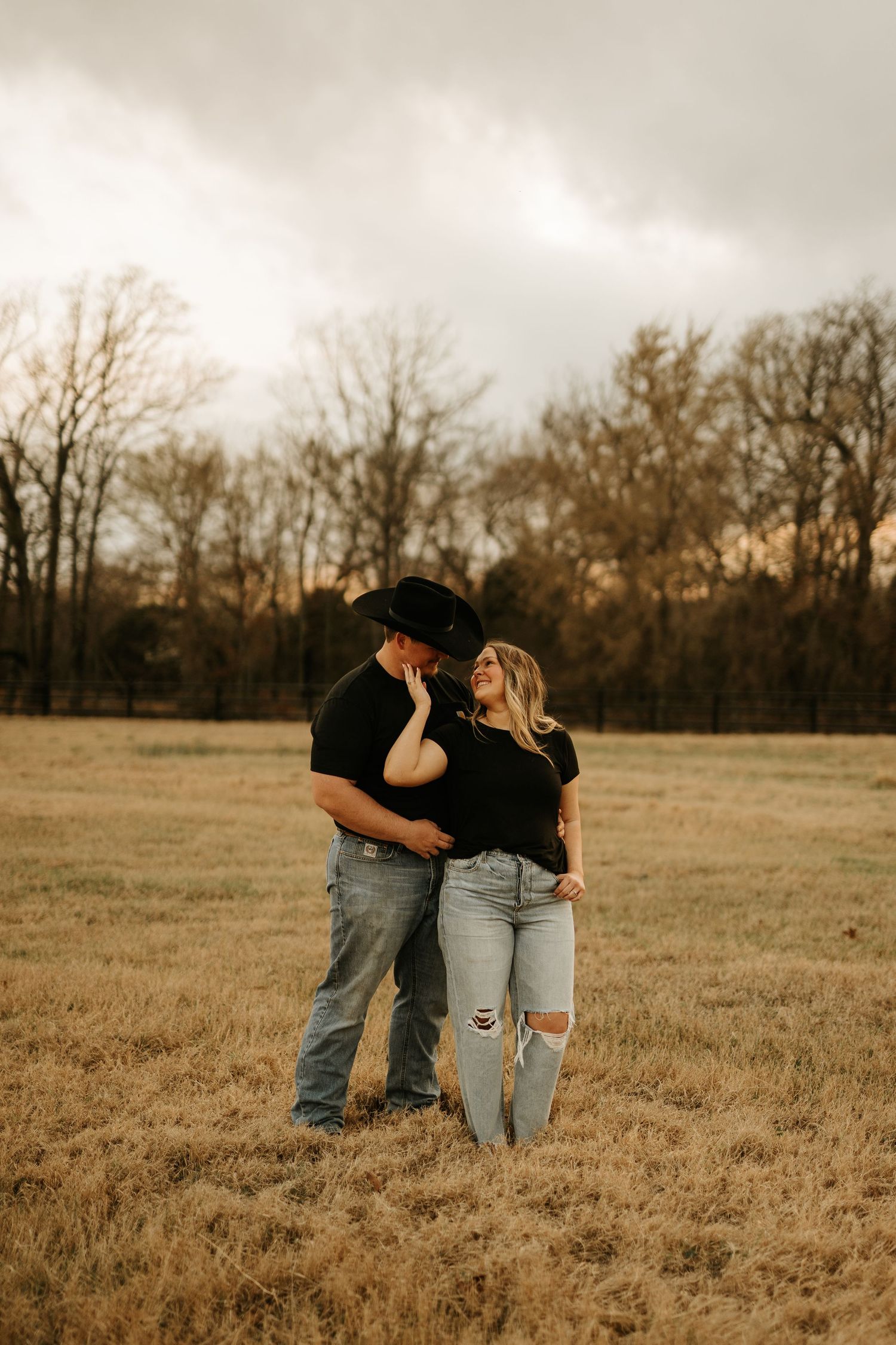 Couple in black tops embracing in golden field, man in cowboy hat, woman in ripped jeans, bare trees and cloudy sky behind them.