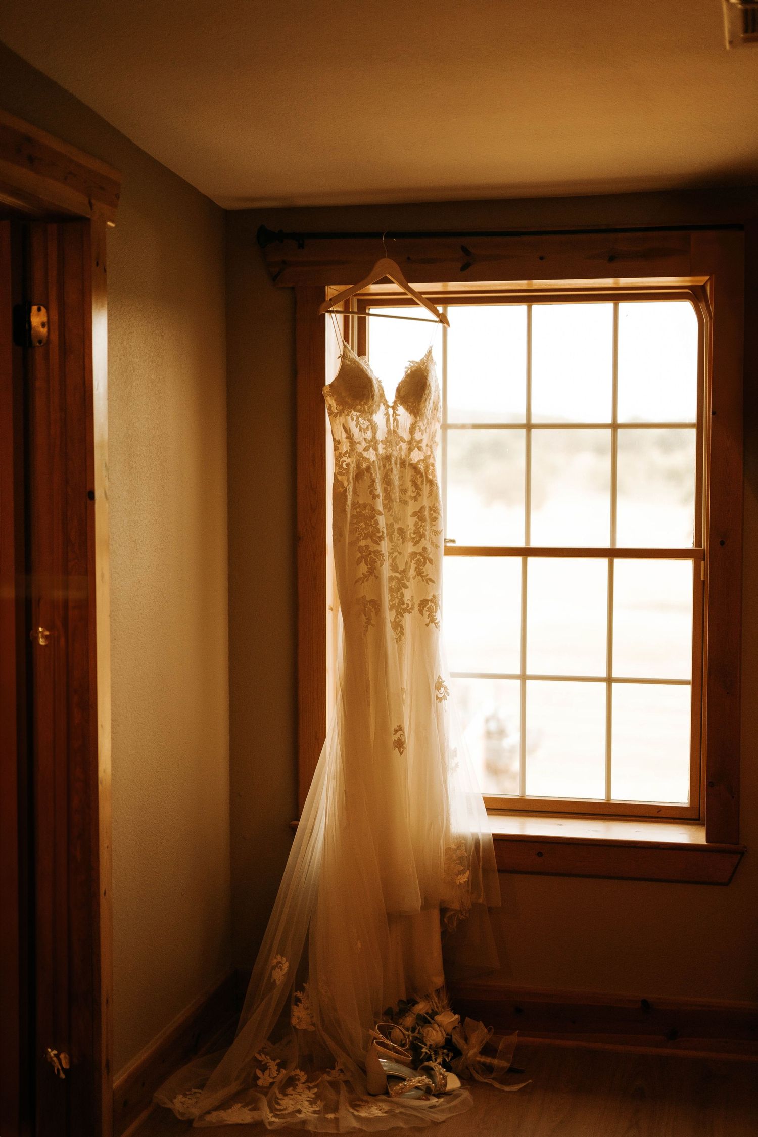 An elegant lace wedding dress hangs by a window in soft, natural light.