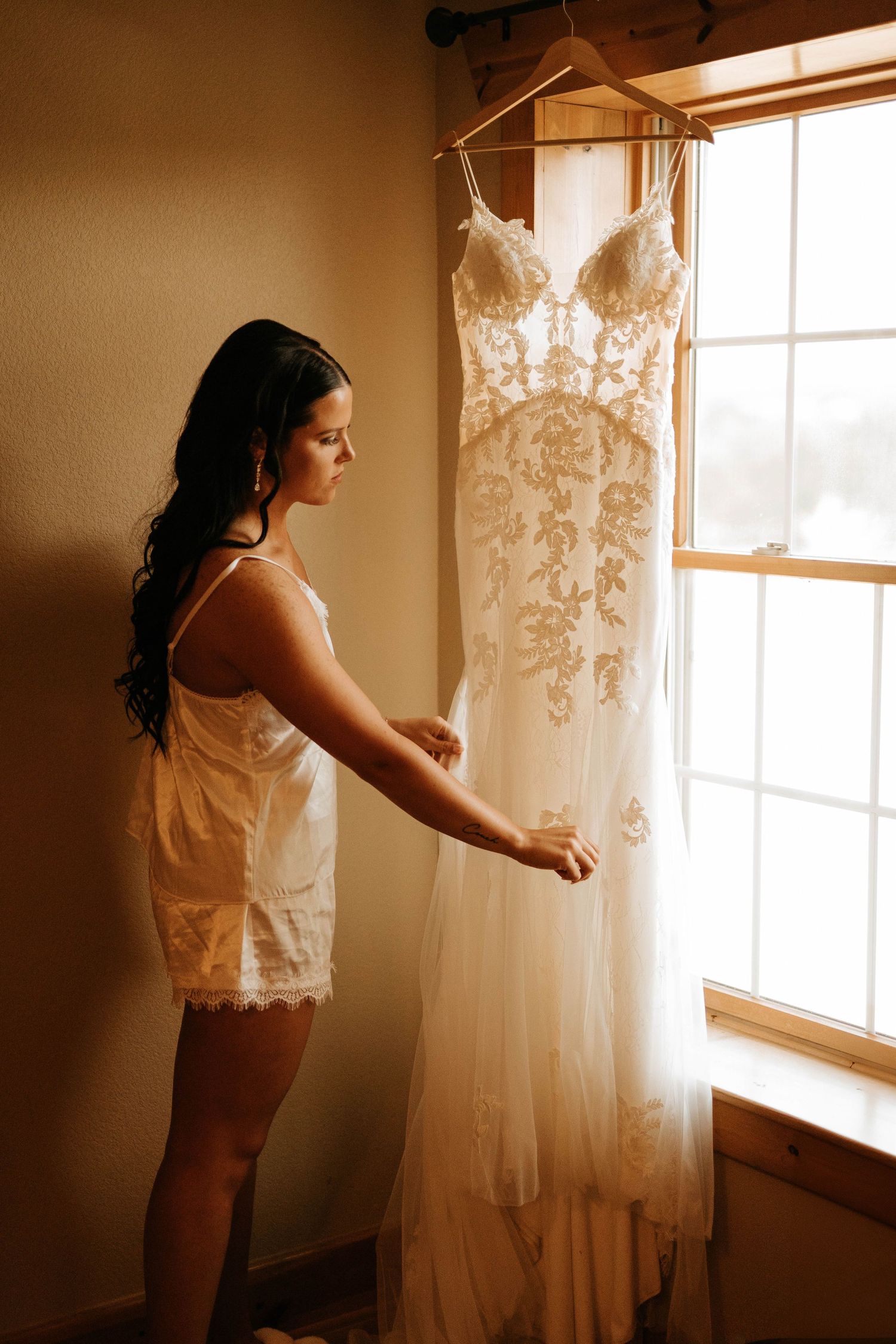 A delicate lace wedding dress hangs by a window as a figure admires it in morning light.