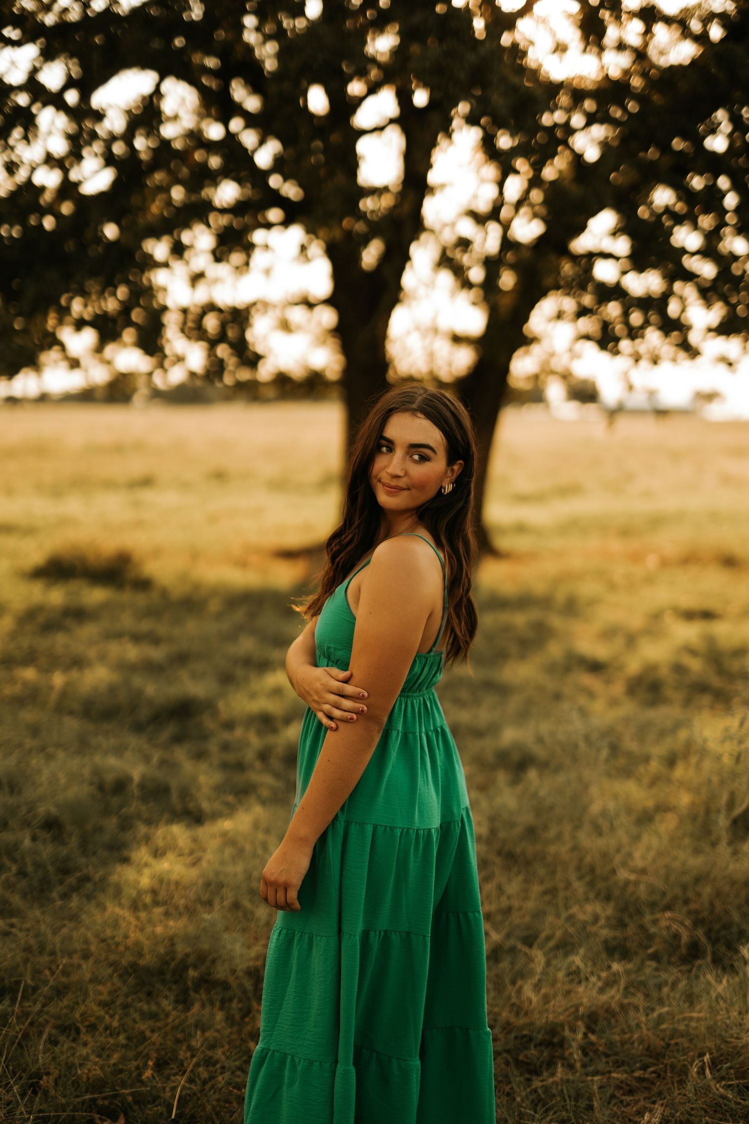 Young woman in green sundress poses gracefully in golden hour sunlight with a large tree backdrop in open field.