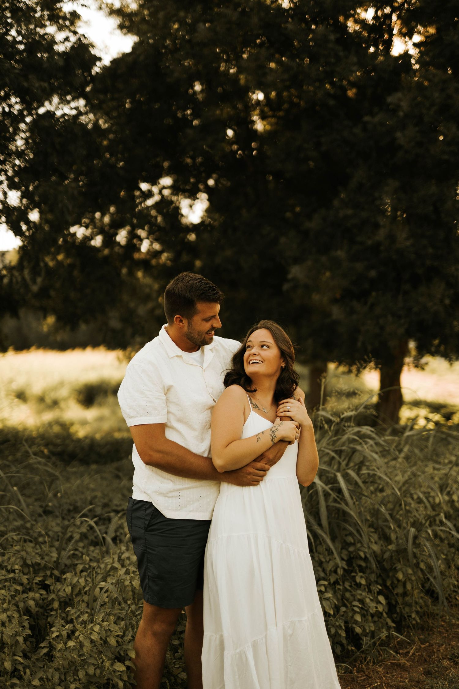 A couple embraces in a sunlit field, the woman in a white dress smiling up at her partner surrounded by lush greenery.