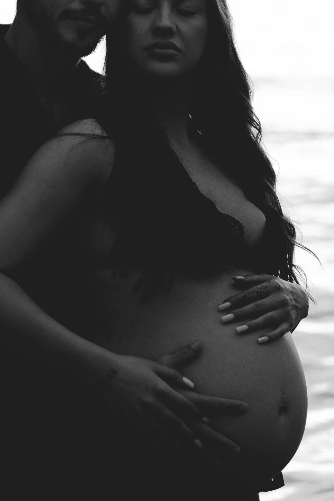 Black and white photo of a pregnant woman cradling her baby bump with her partner embracing her from behind near water.