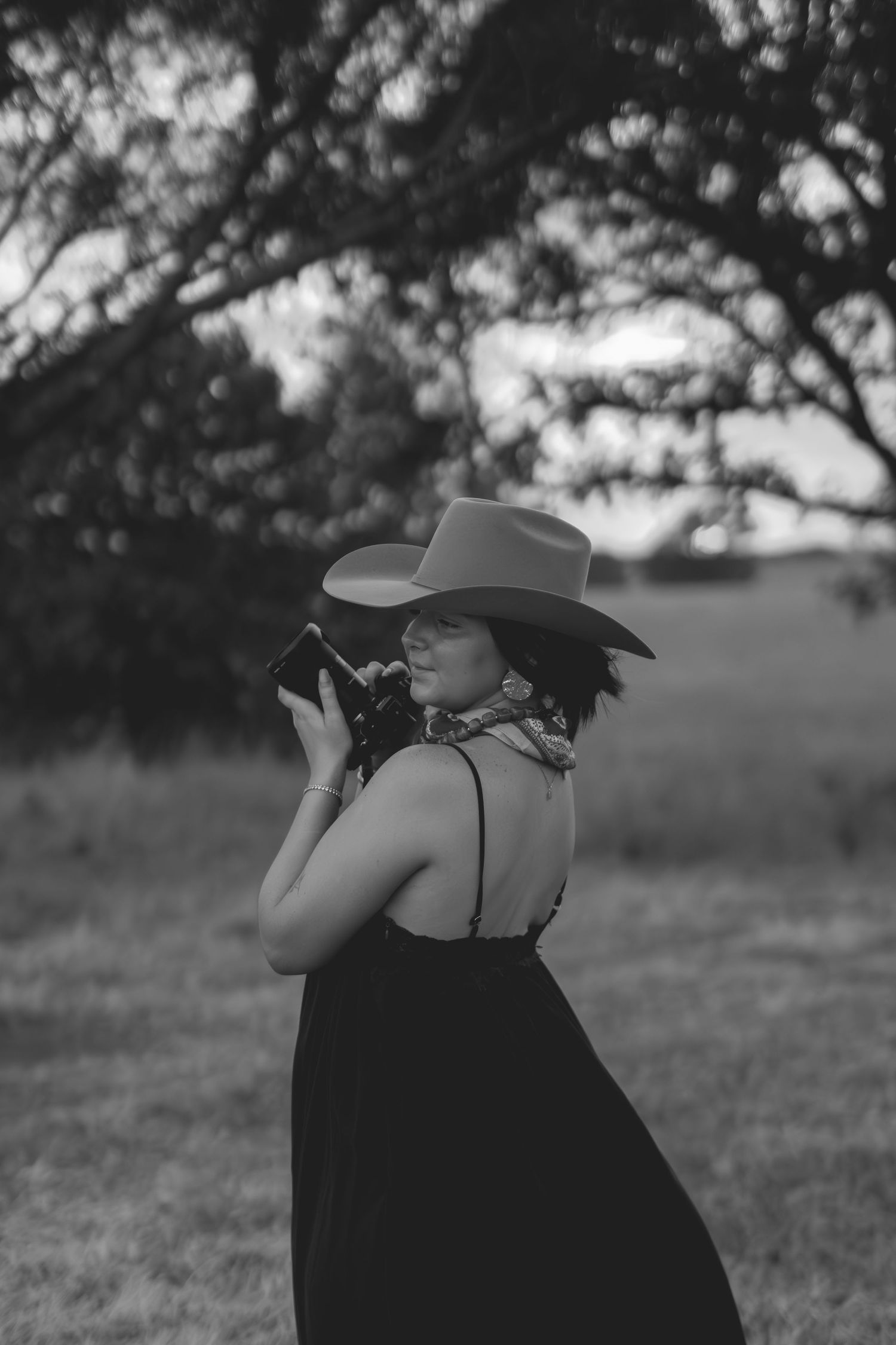 Black and white photo of a woman in a cowboy hat and sundress holding a camera outdoors near a tree.