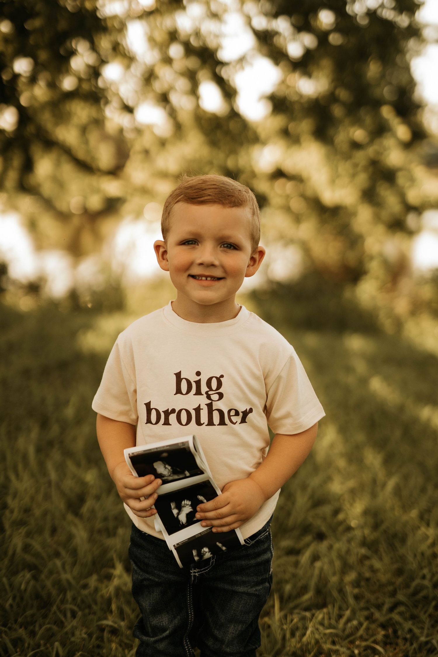 Young boy wearing a 'big brother' t-shirt holds ultrasound photos while smiling in golden outdoor lighting.