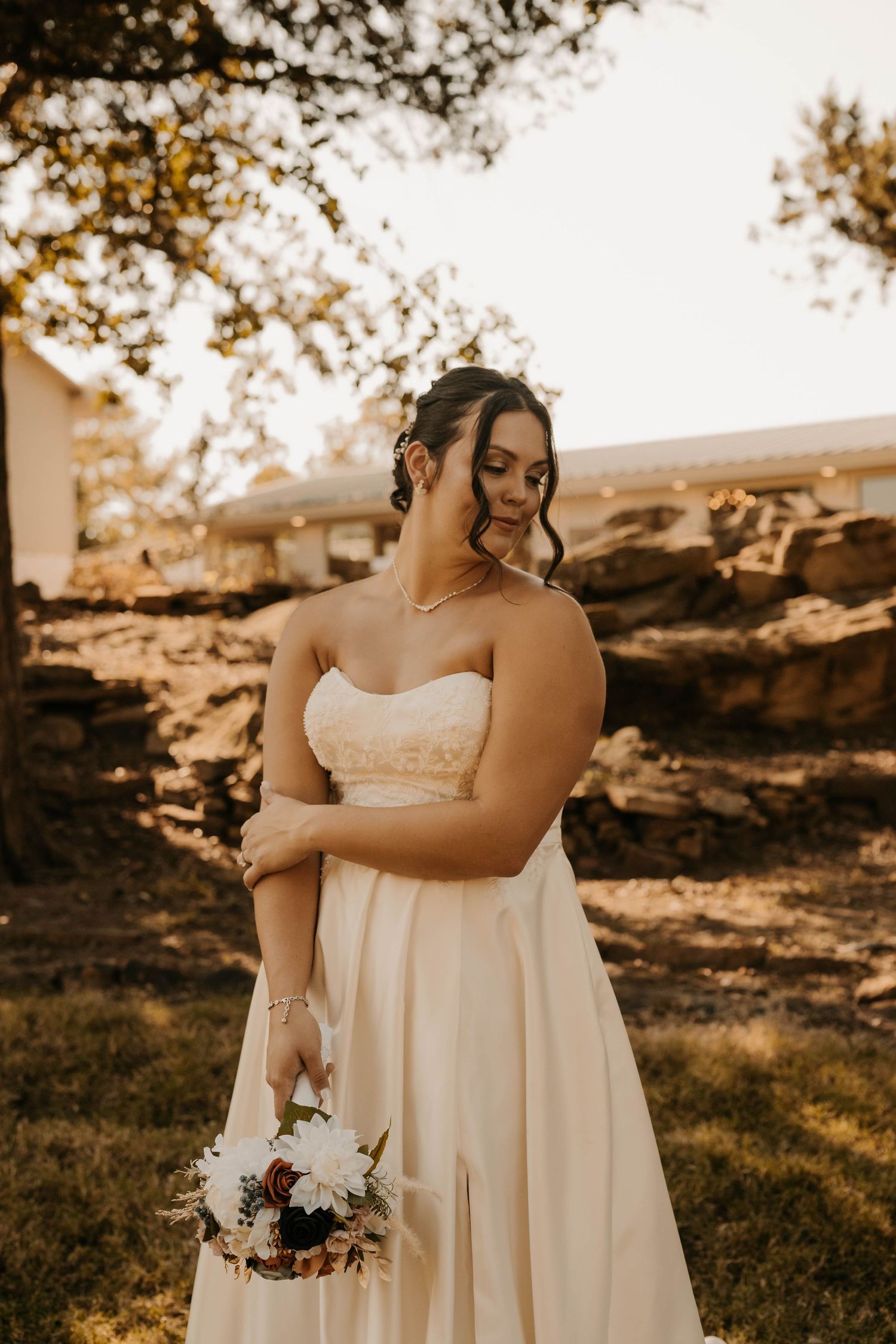 A bride in a strapless white gown holds a bouquet while standing in warm autumn light.