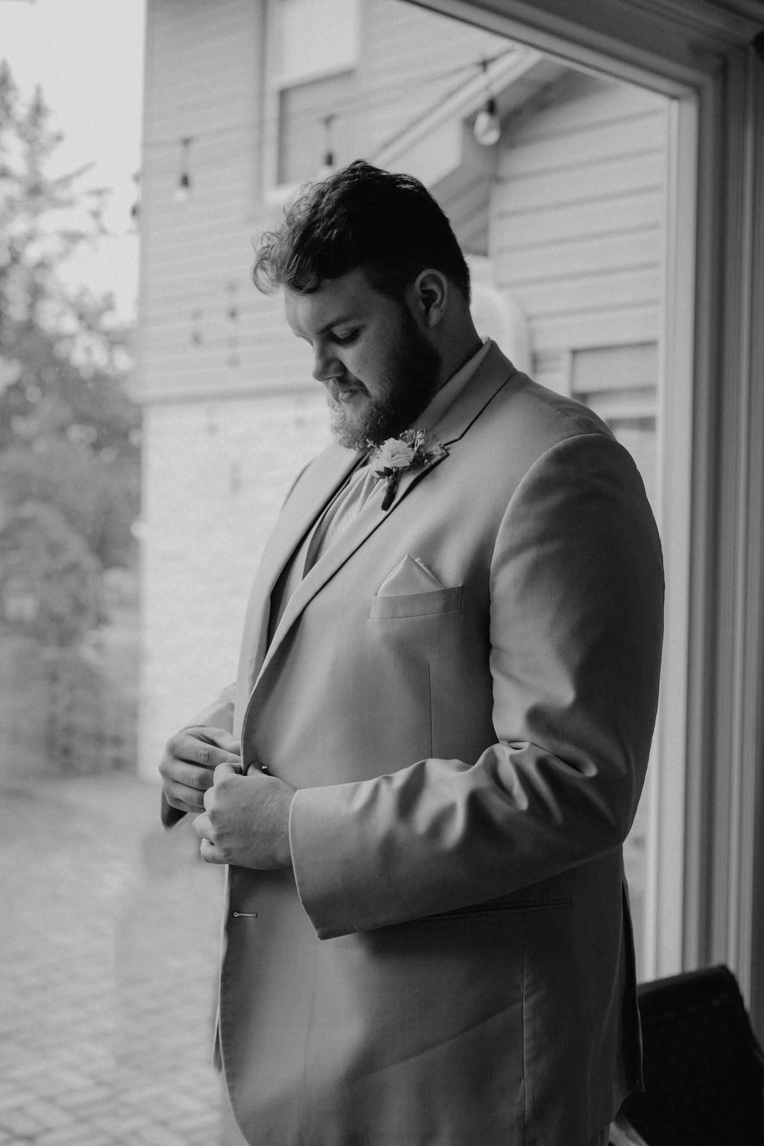 A groom in a light colored suit adjusts his bowtie by a window.