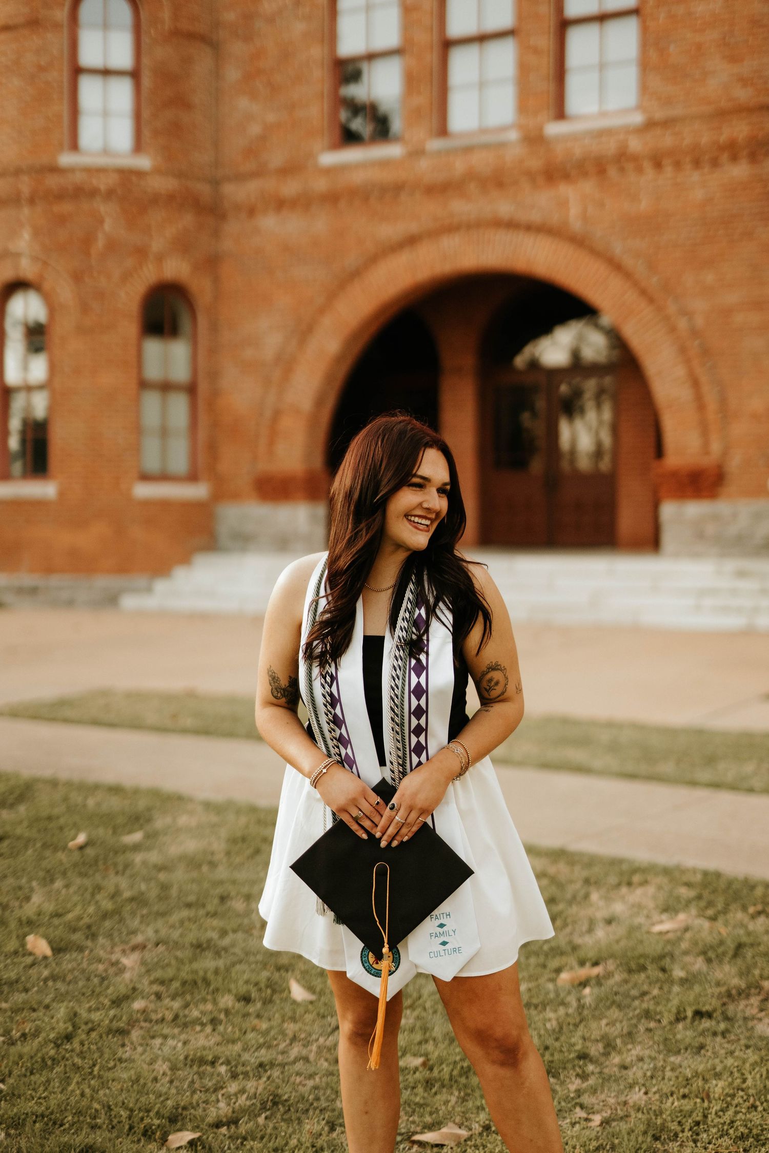 Graduate smiling while holding diploma in front of ornate brick building with arched entrance.