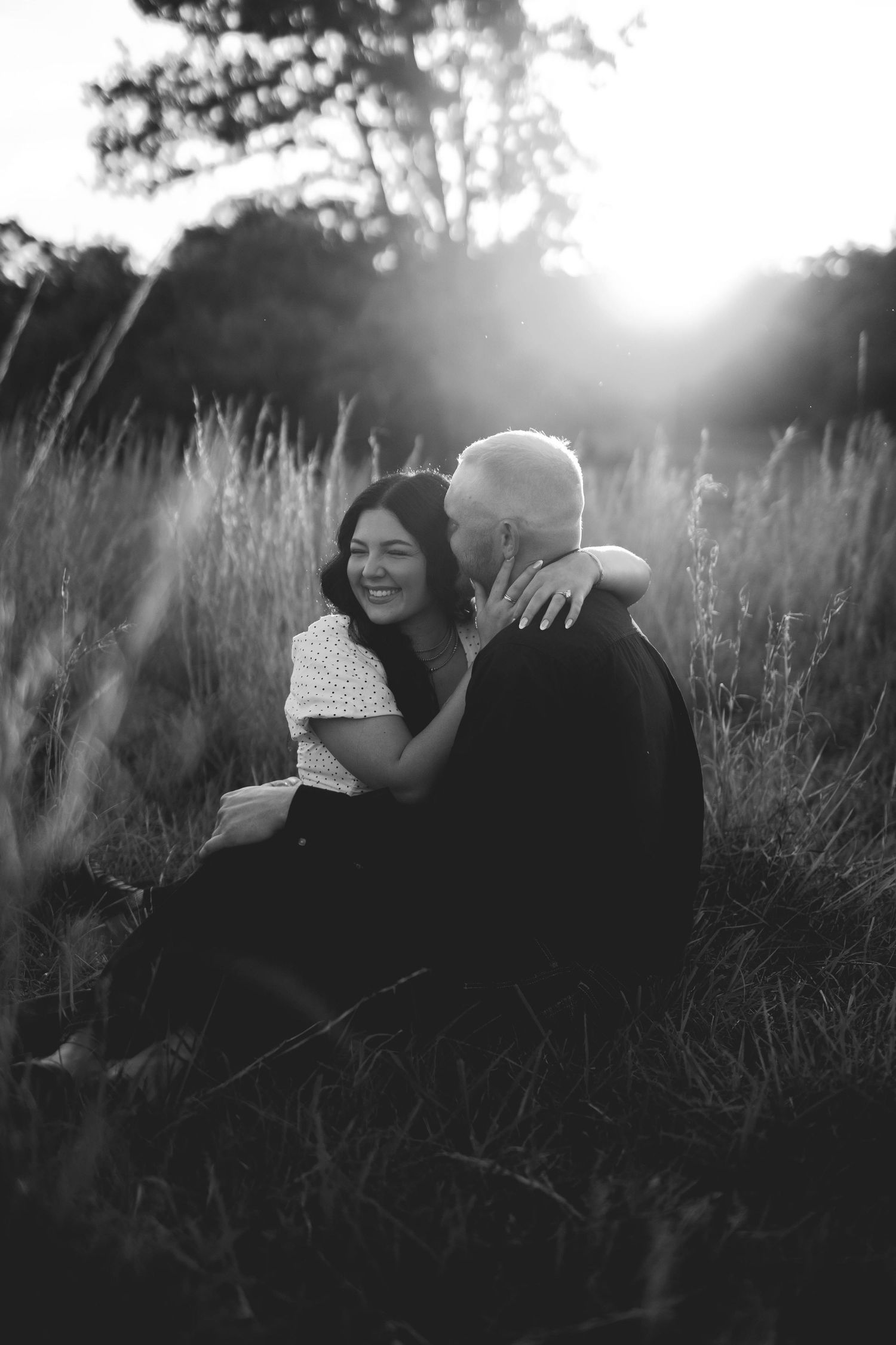 Black and white portrait of a couple embracing in tall grass during sunset.