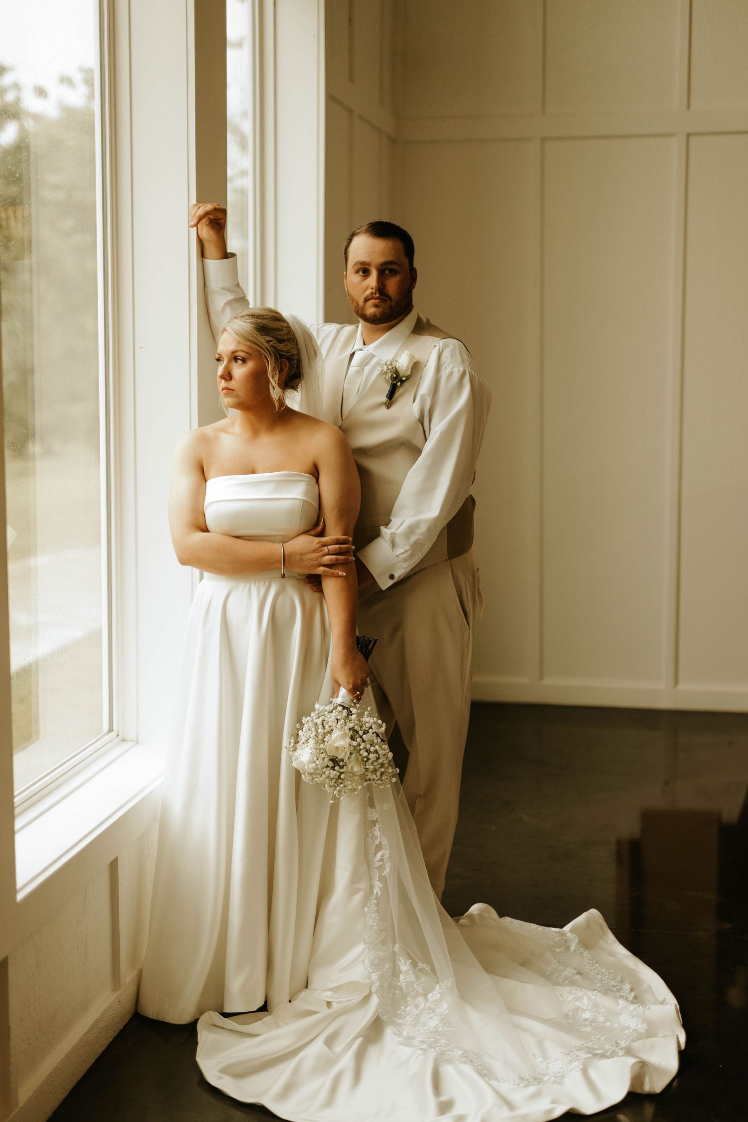 Couple poses by large window in wedding attire with long train dress.