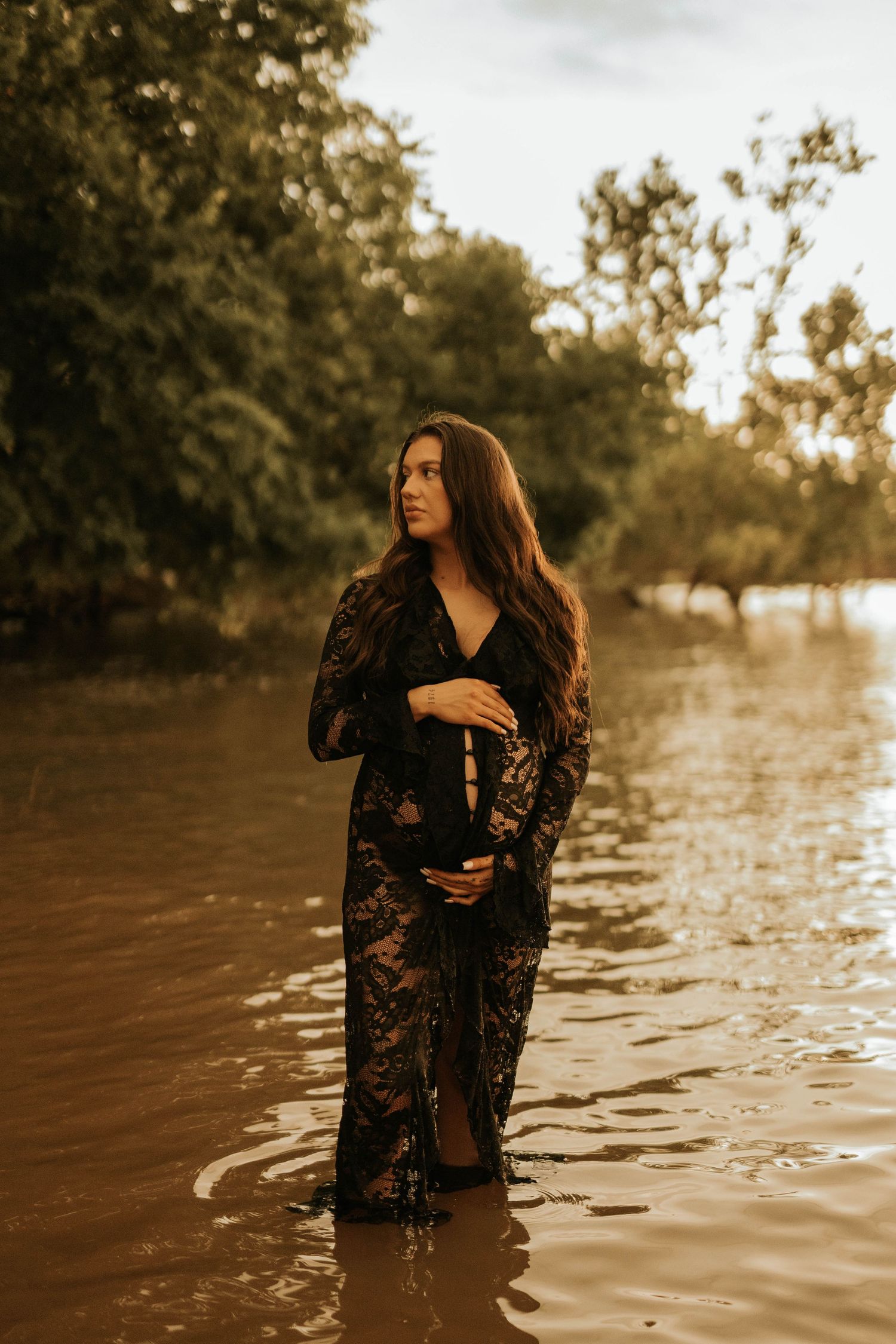 Pregnant woman in black lace gown standing in shallow water at golden hour, cradling baby bump.