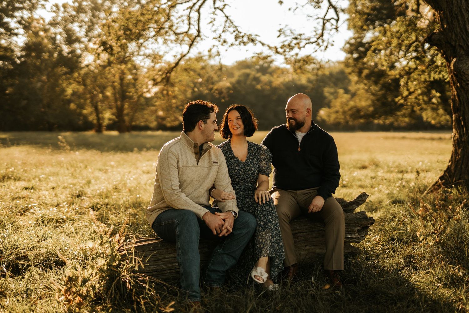 Three people sitting on a log in a sunlit meadow, sharing a warm conversation during golden hour.