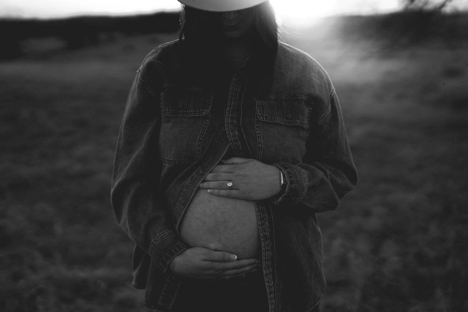 Black and white photo of a pregnant woman in a denim jacket cradling her baby bump in a field at sunset.