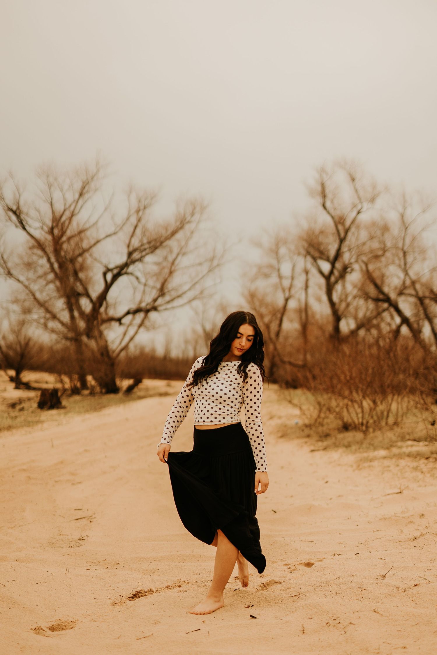 Woman in polka dot top and black skirt walking barefoot on sandy desert path with bare trees in background.
