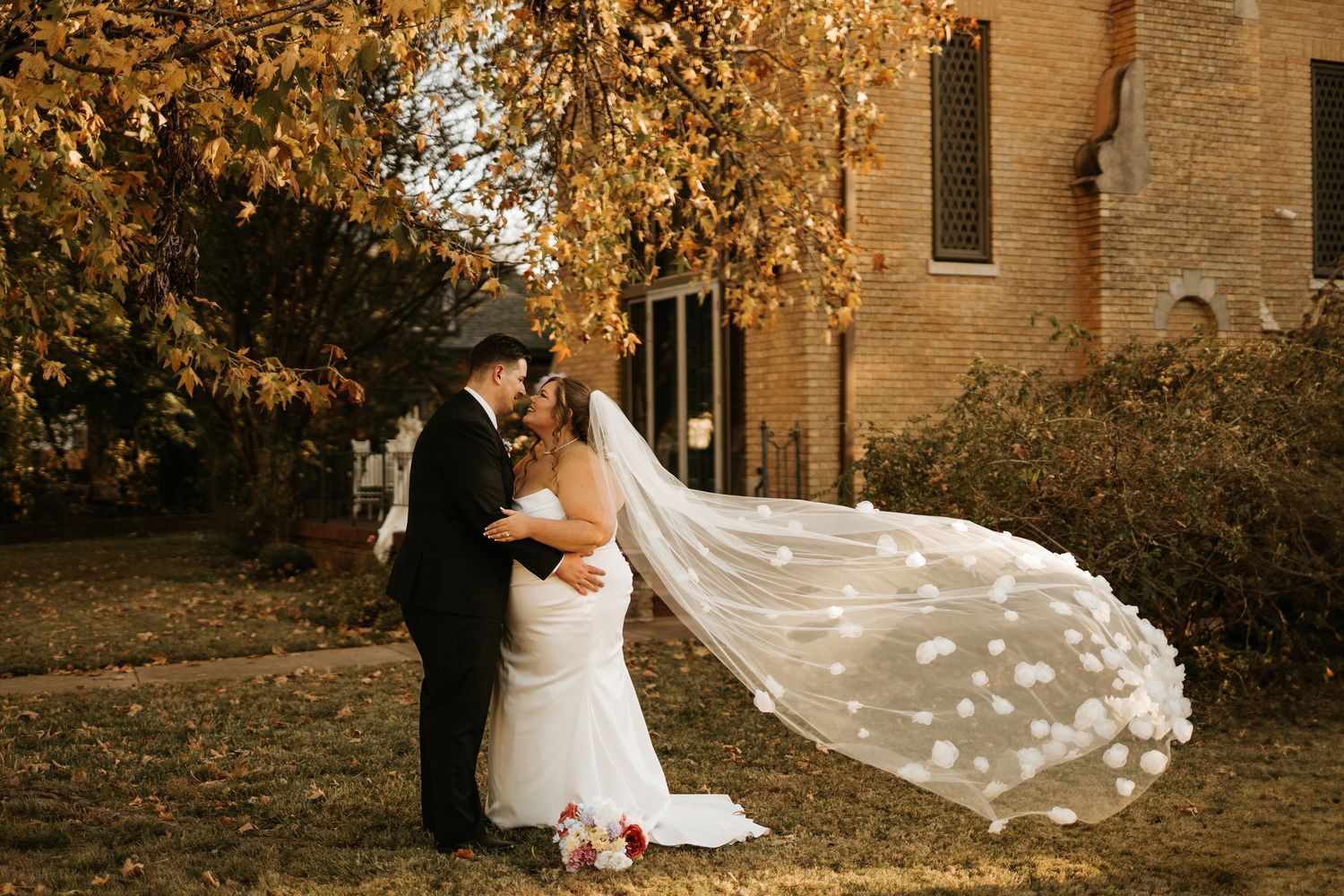 Bride and groom sharing a romantic kiss outdoors in autumn, with a flowing floral veil billowing in the wind near a brick building.