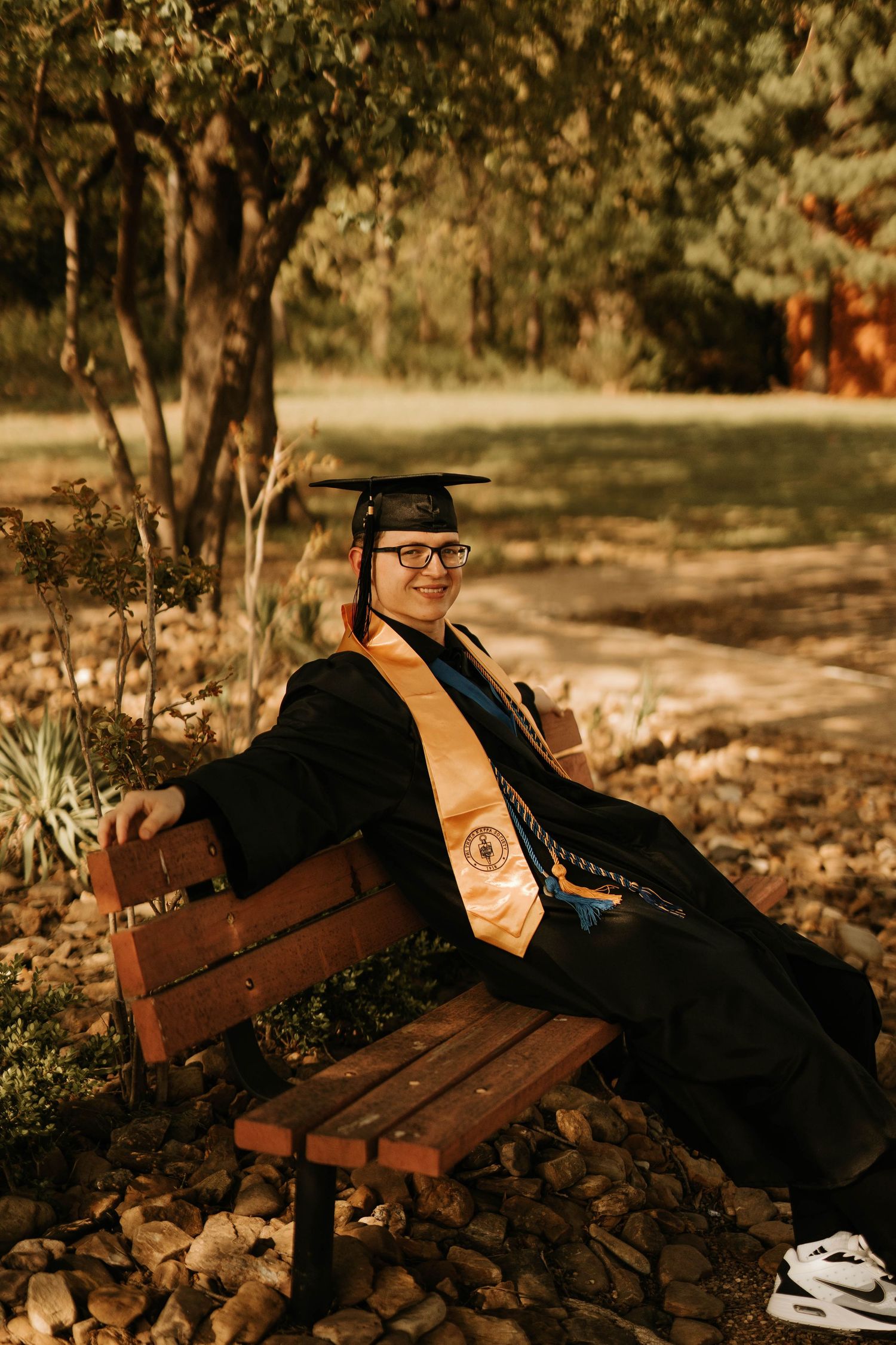 Graduate in black gown and orange stole relaxes on wooden bench in autumn setting.
