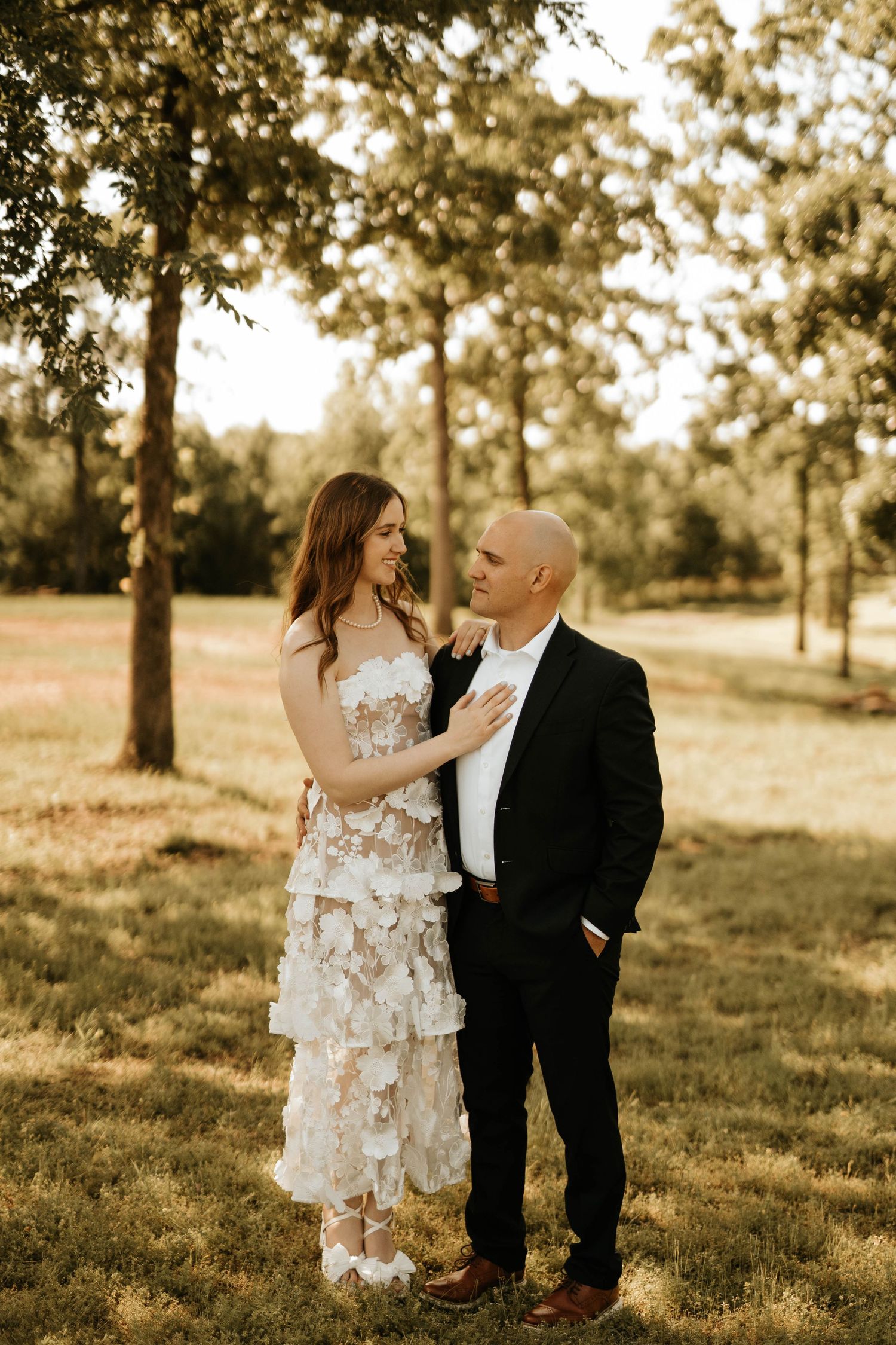 Newlyweds share intimate moment in sunlit garden, bride in tiered dress and groom in classic black suit.