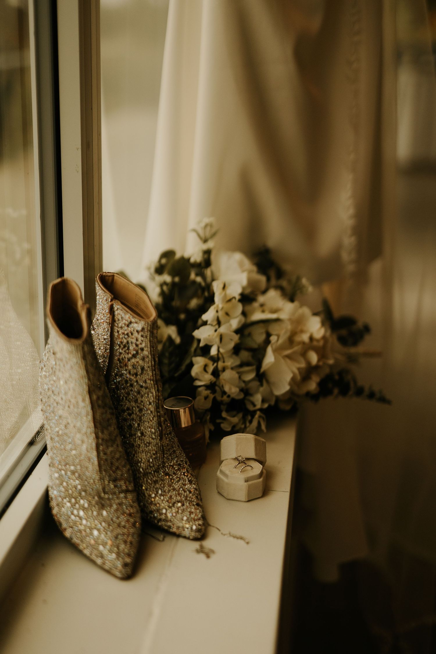 Glittery cowboy boots beside a floral bouquet and ring box on a windowsill for a wedding.