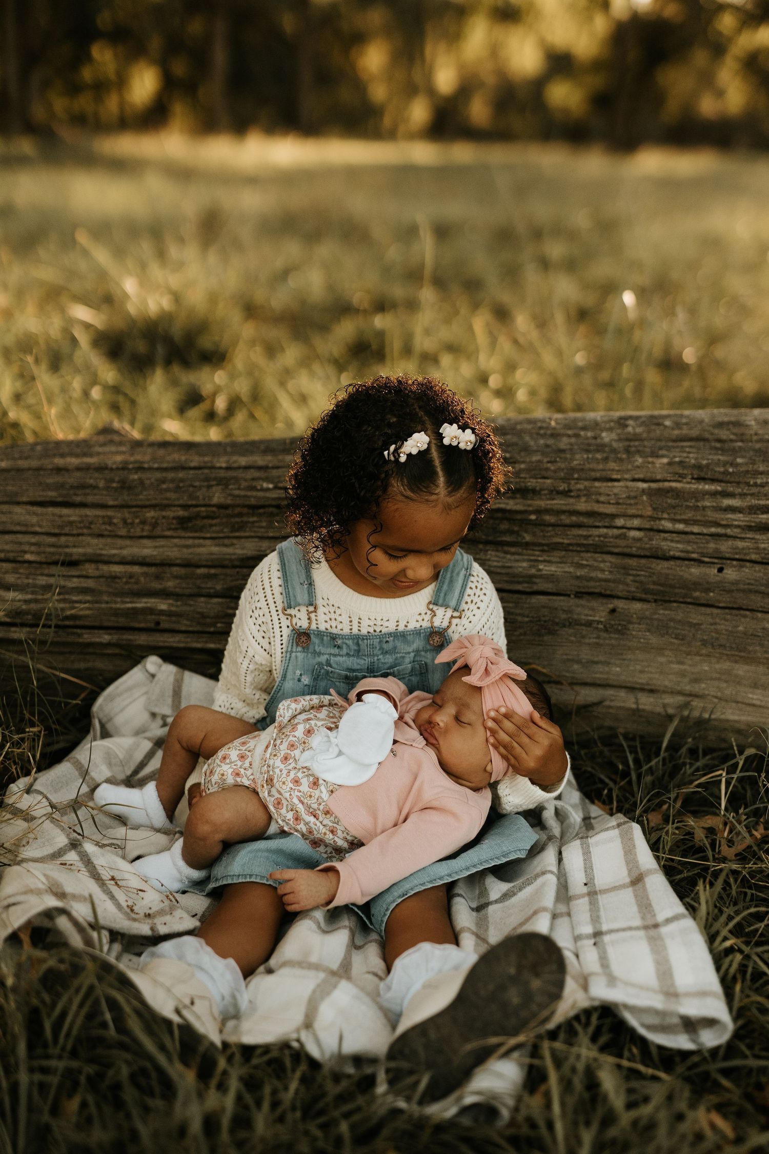 Young girl with curly hair holds sleeping newborn baby outdoors on plaid blanket near rustic wooden fence.