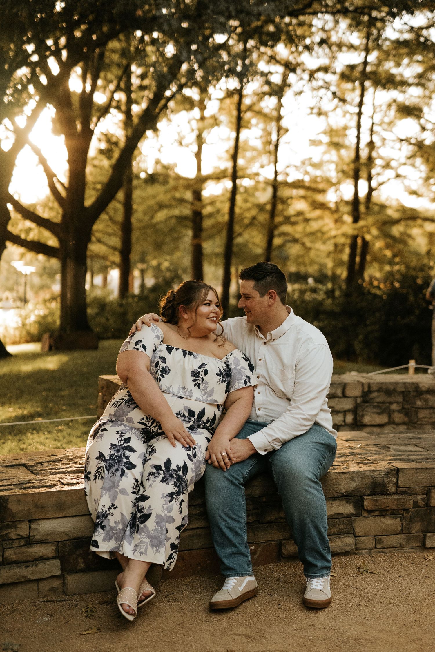 Couple sitting on stone bench at golden hour, woman in floral dress and man in white shirt sharing a romantic moment outdoors.