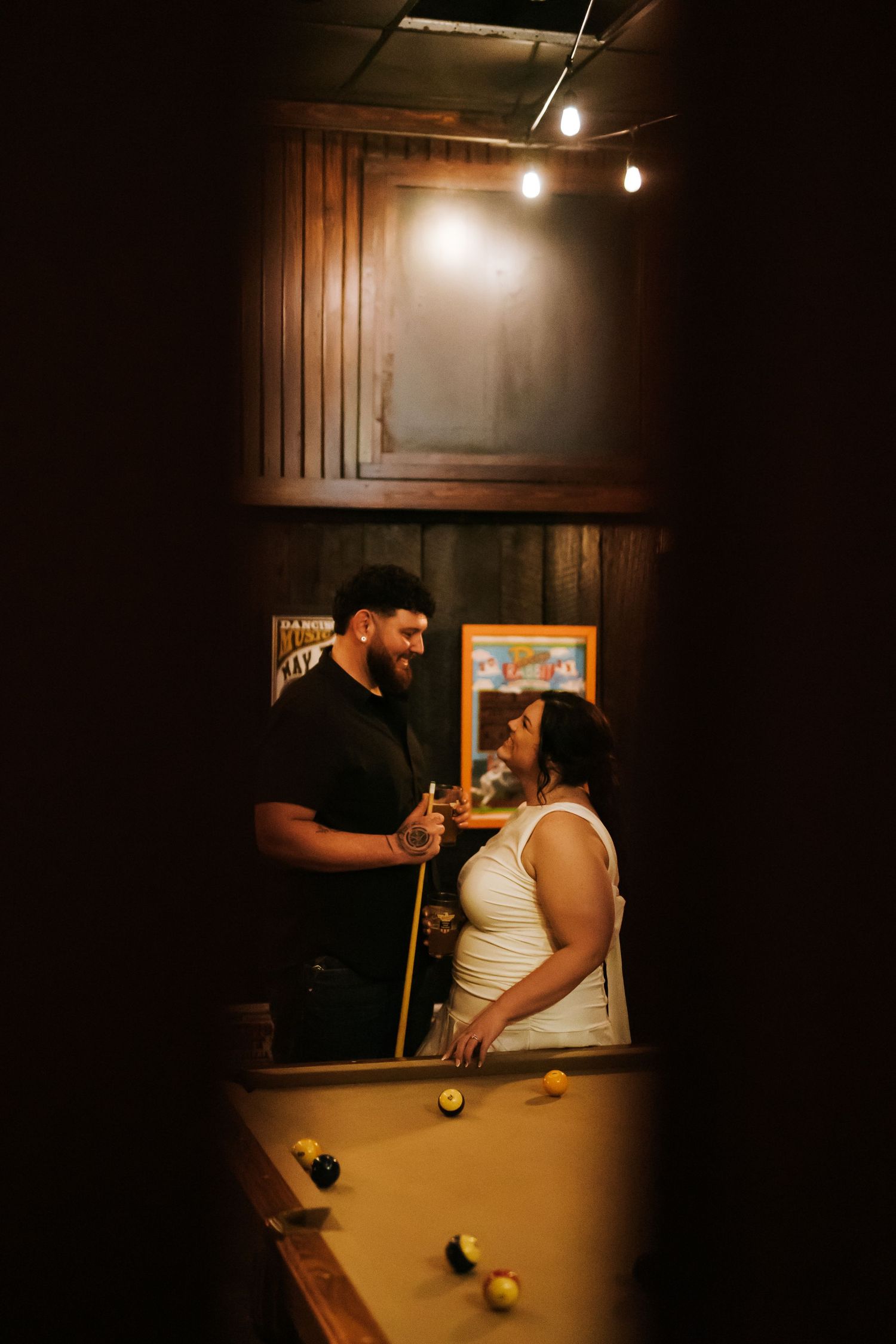 Couple sharing a romantic moment over a pool table in a warmly lit bar with string lights and wood-paneled walls.