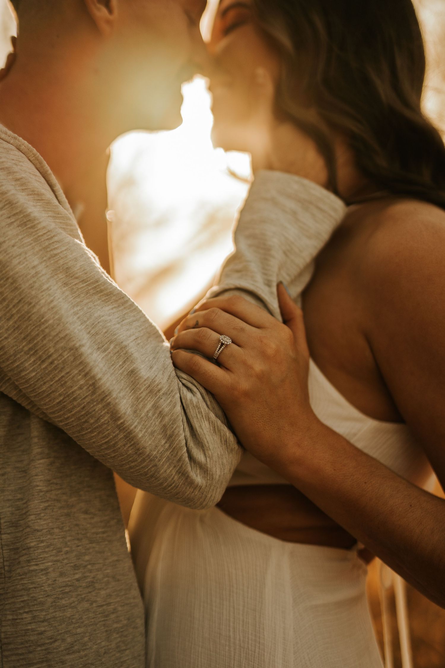 Couple embracing in golden sunlight, woman wearing engagement ring in white dress, romantic outdoor photo.