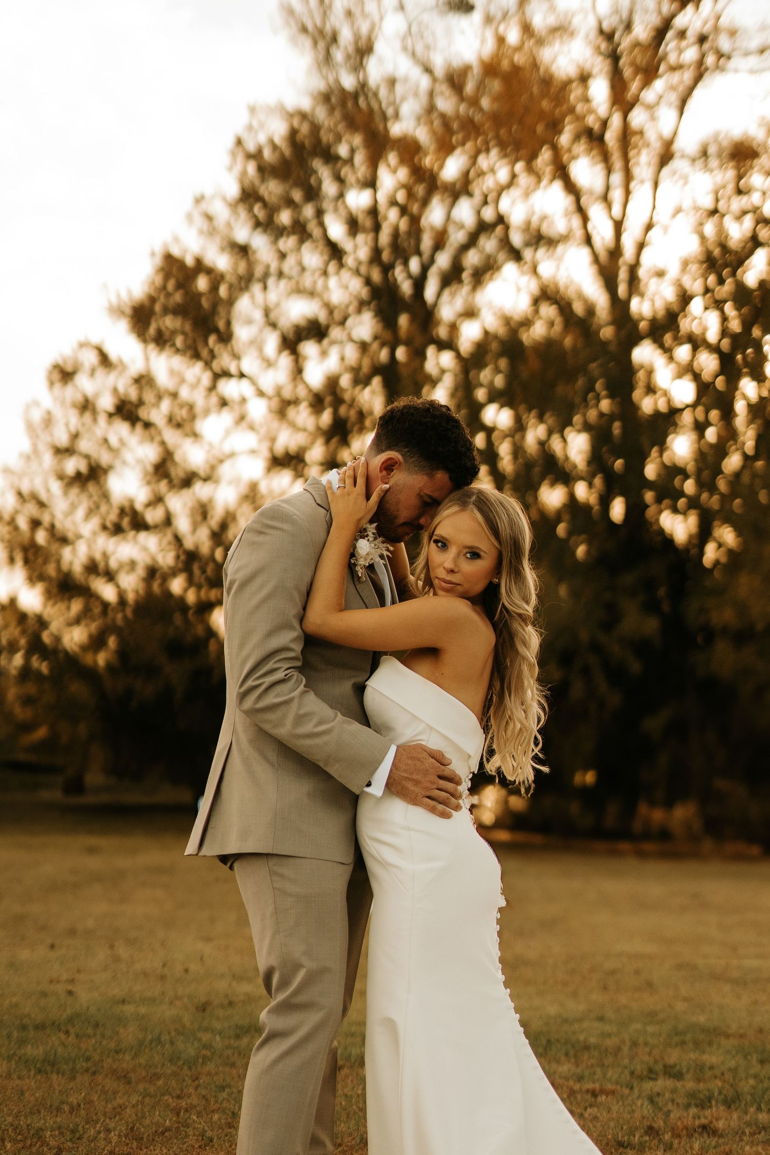 Bride in white gown and groom in tan suit embrace outdoors during golden hour wedding portrait.