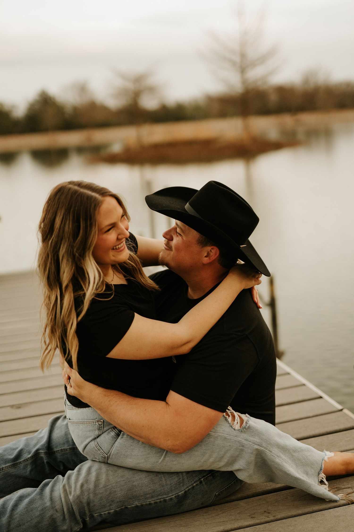 Couple sitting on a dock embracing and smiling, man wearing a black cowboy hat, with a lake in the background.
