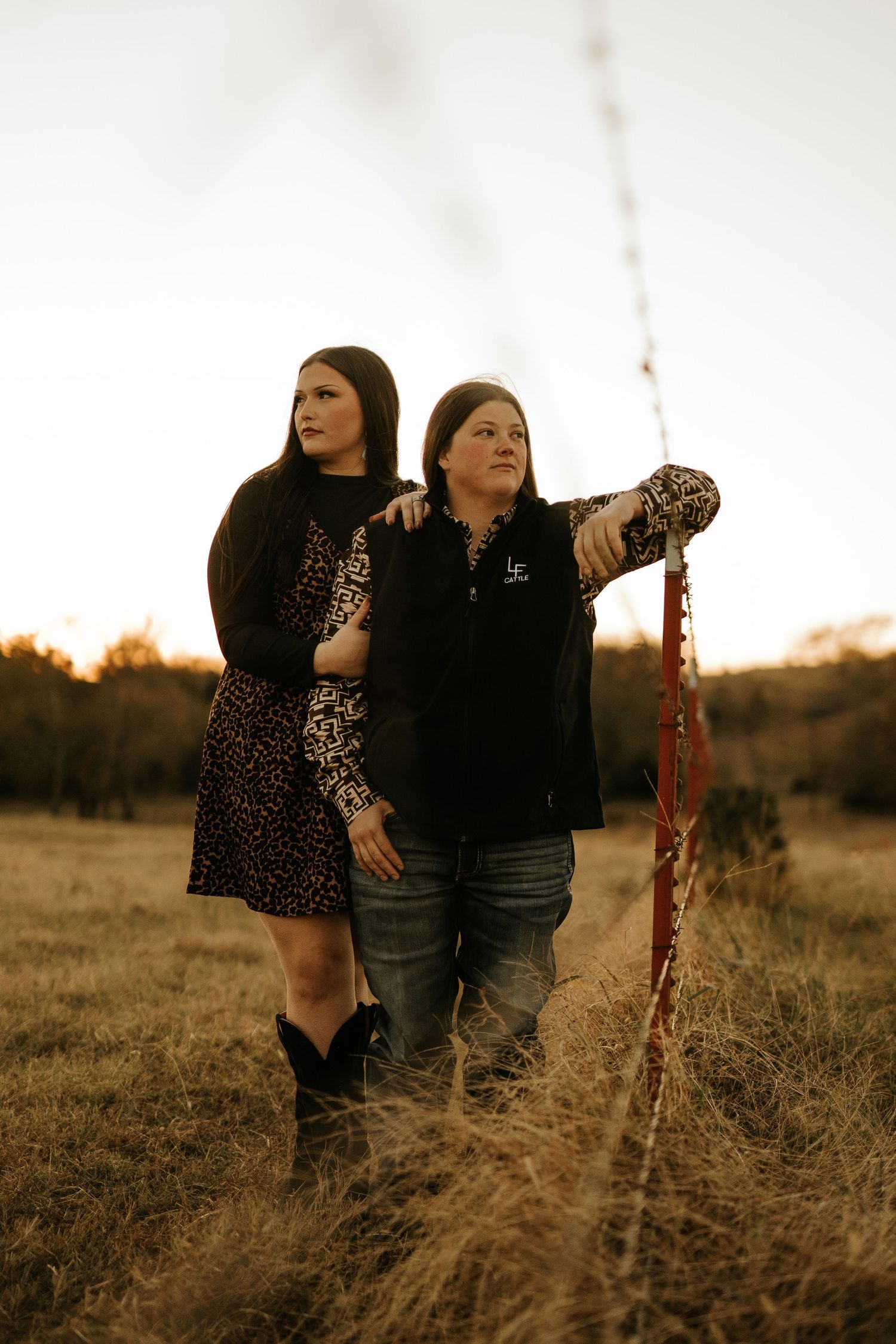 Two women pose together in a golden field at sunset, one in leopard print dress with boots, other in black jacket and jeans.