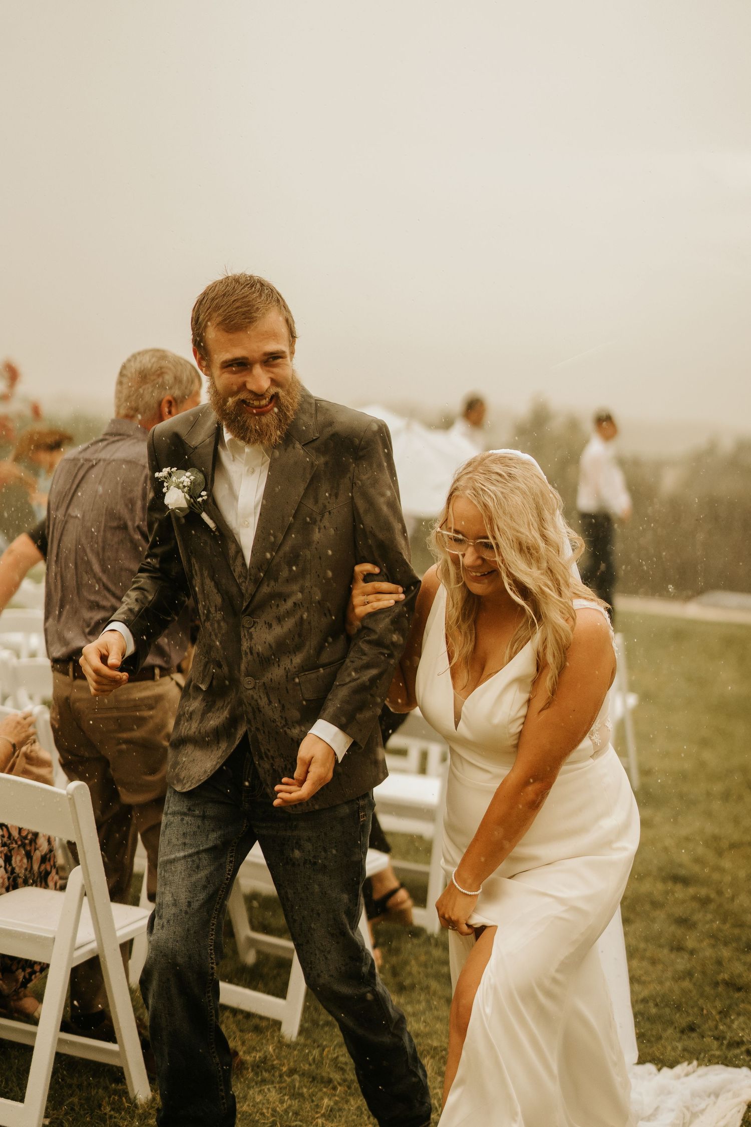 Bride and groom laughing in the rain during outdoor wedding ceremony recessional, bride in white gown, groom in gray suit.