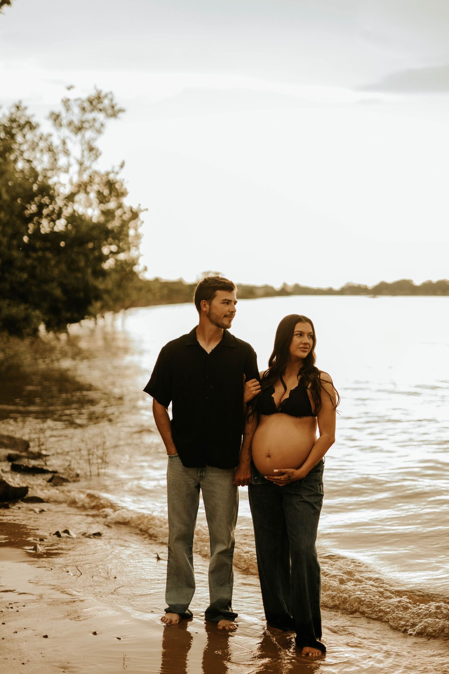 Pregnant couple standing barefoot on a sandy beach shore at sunset, woman cradling baby bump in jeans and bikini top.