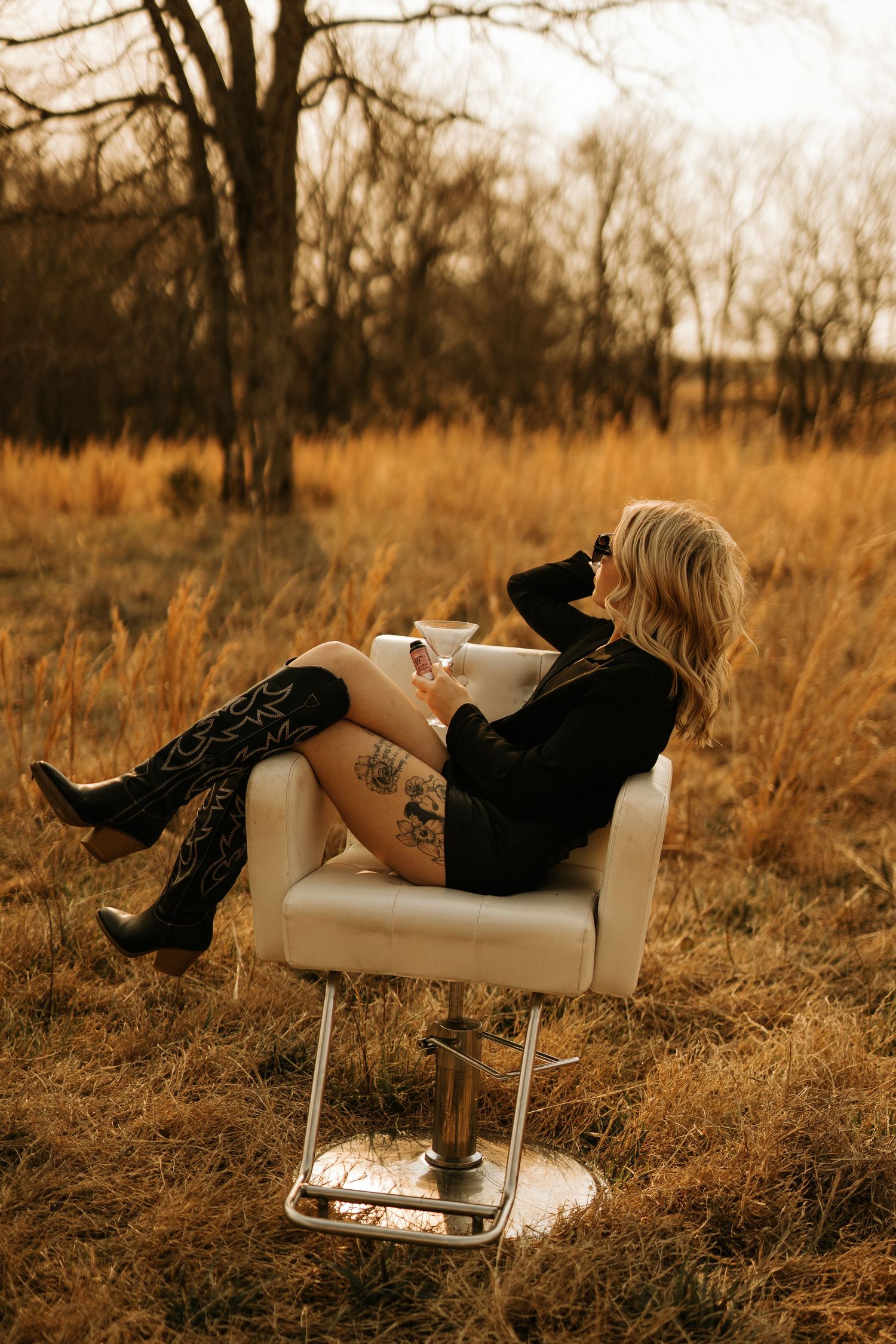 Blonde woman with tattoos in black dress and cowboy boots sitting in salon chair outdoors in golden field.