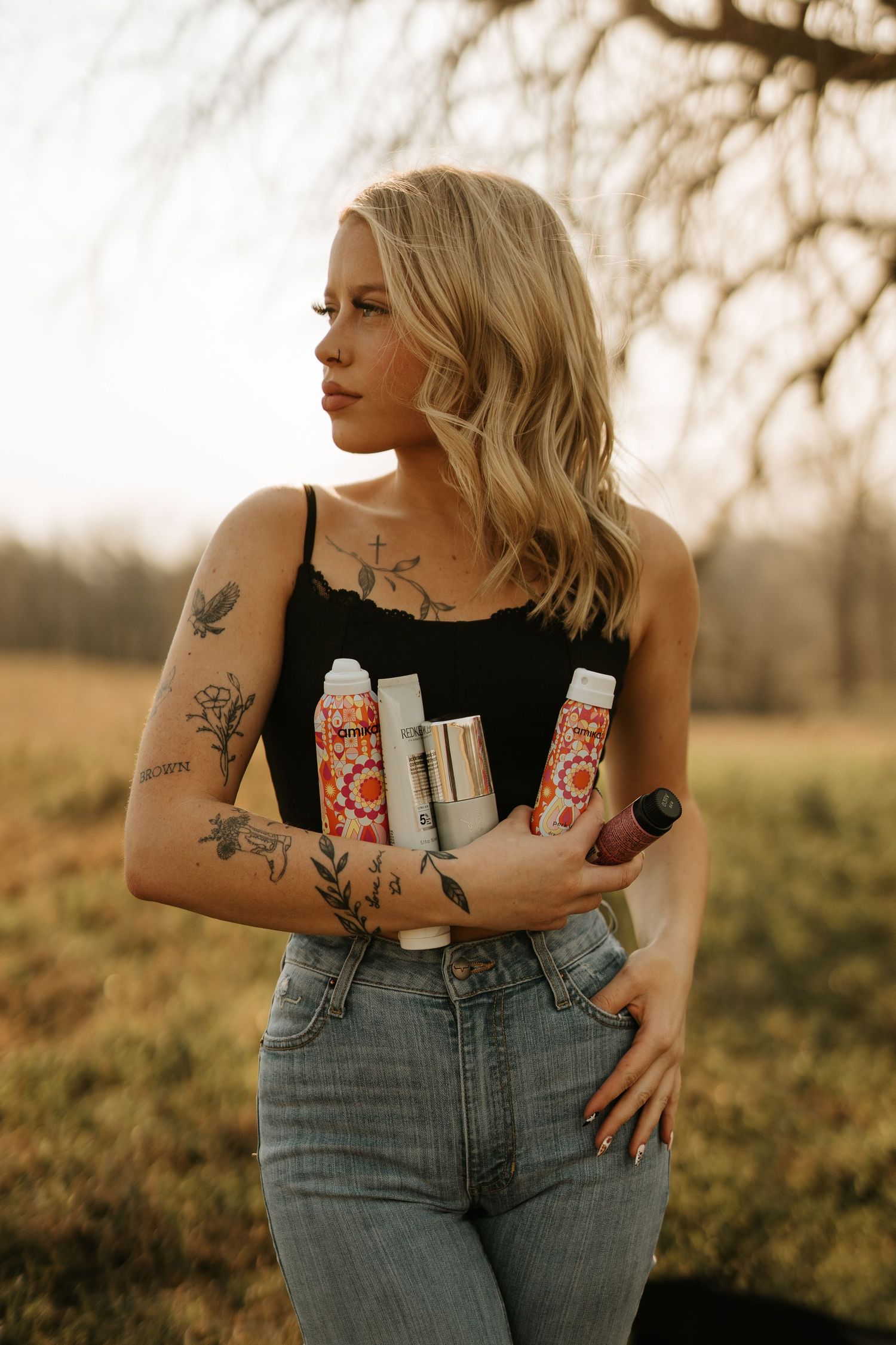 Tattooed blonde woman in black top and jeans holding hair care products outdoors in a golden field setting.