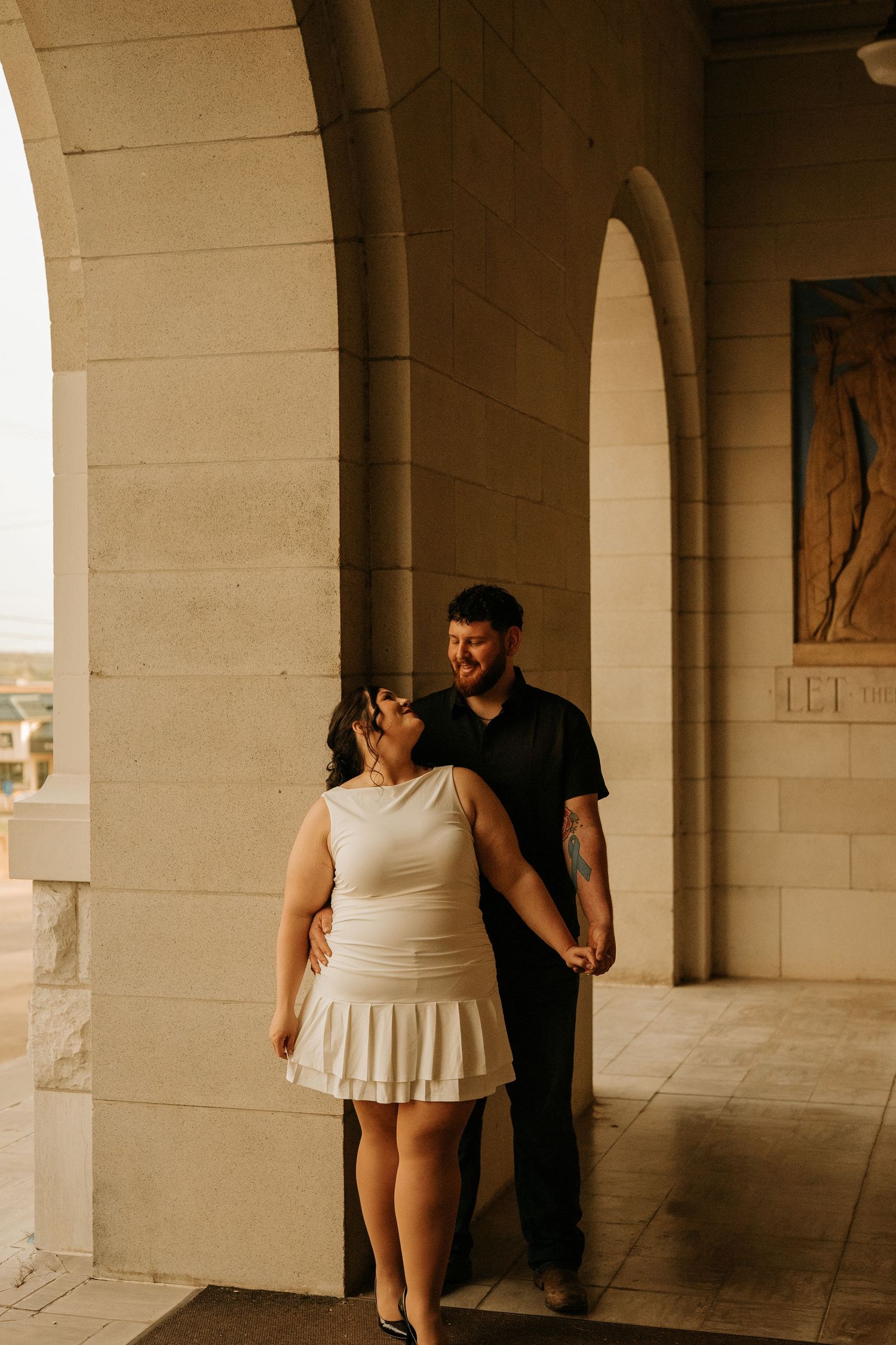 Couple holding hands and smiling at each other under stone arches, woman in white dress, man in black shirt.