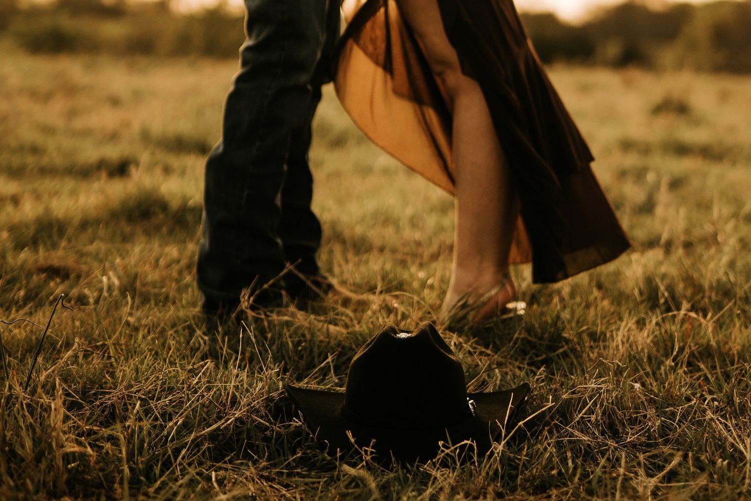 Couple sharing a romantic kiss in a golden field at sunset with a black hat resting on the grass.
