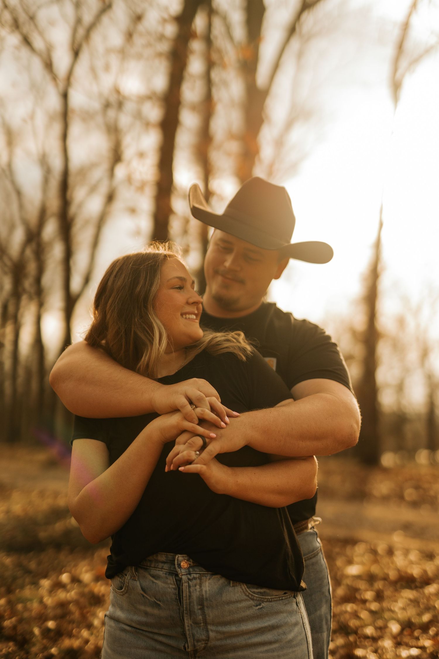 Couple embracing in autumn forest at golden hour, man in black cowboy hat and shirt, woman smiling in black top and jeans.