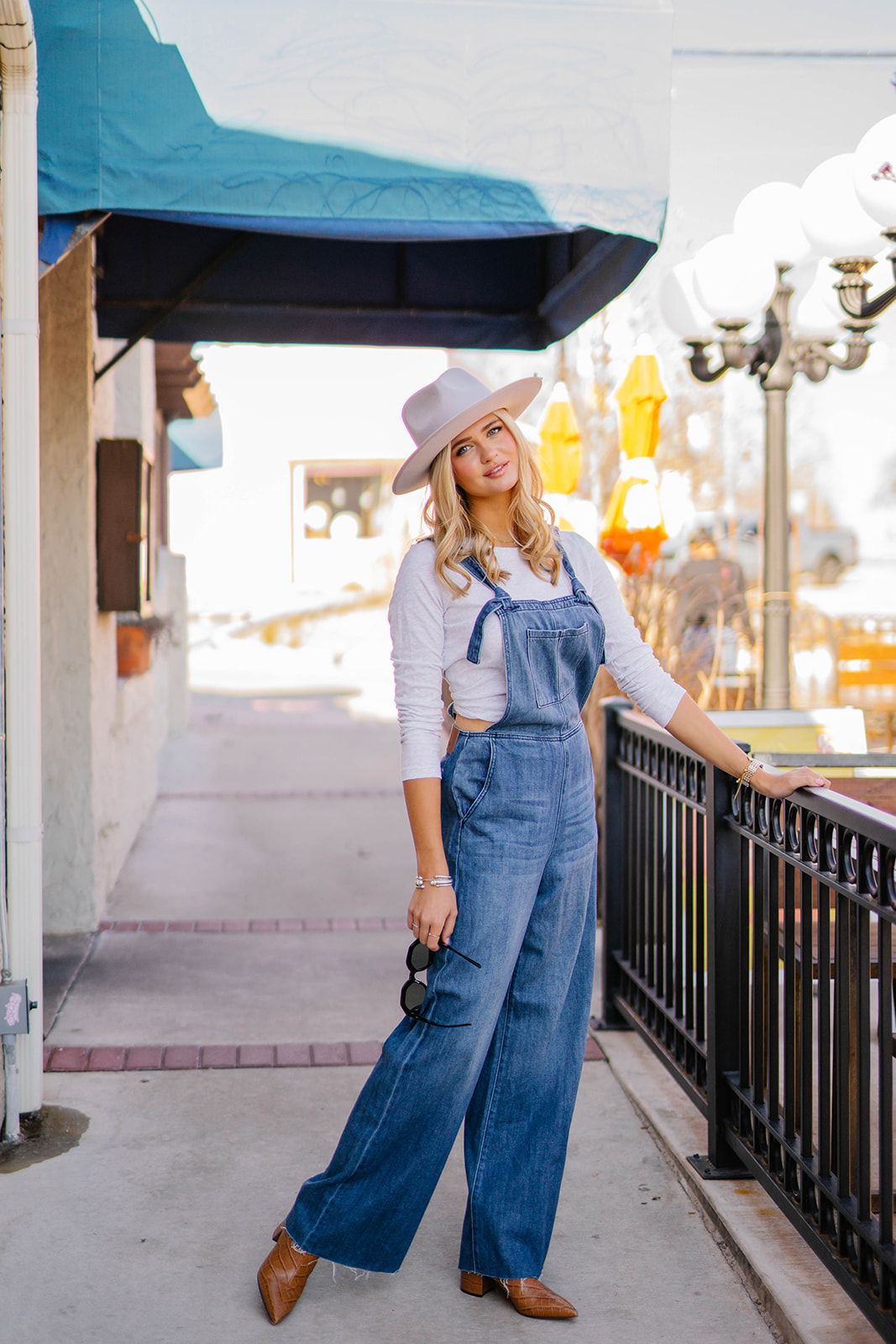 Stylish woman in denim overalls, white top and hat poses casually against storefront railing.