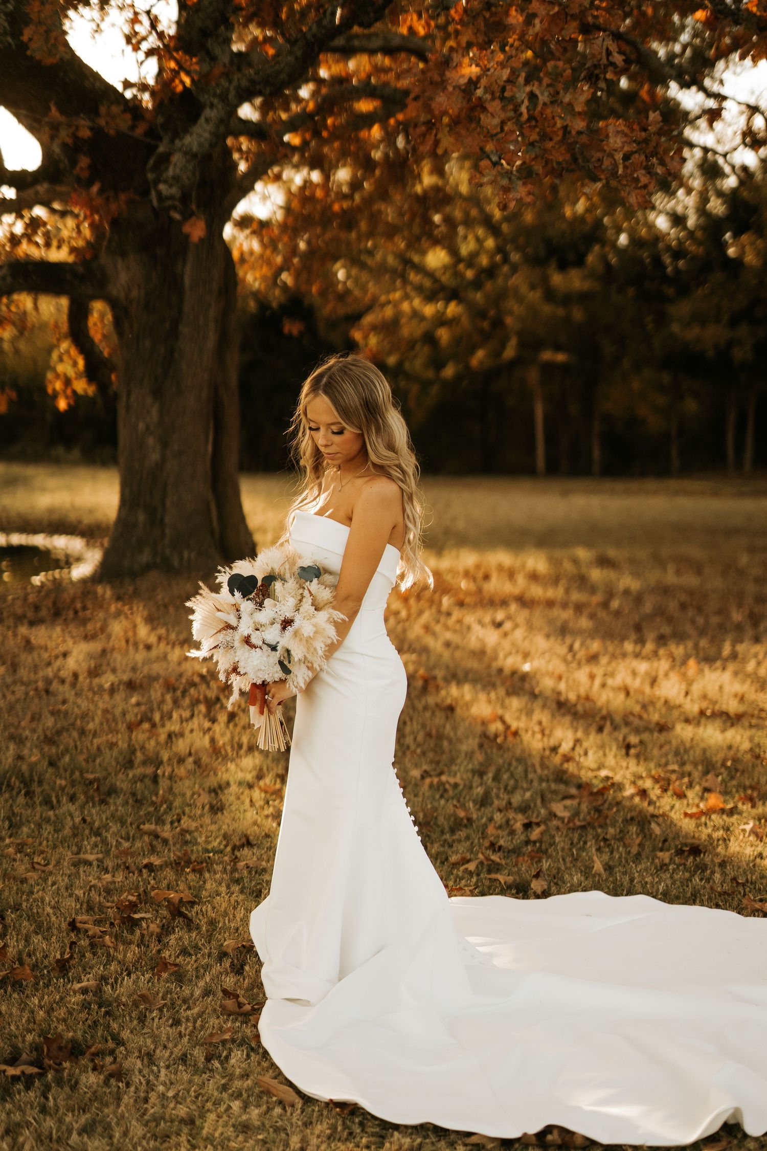 Bride in strapless white mermaid gown holding bouquet under autumn tree with golden foliage in outdoor setting.