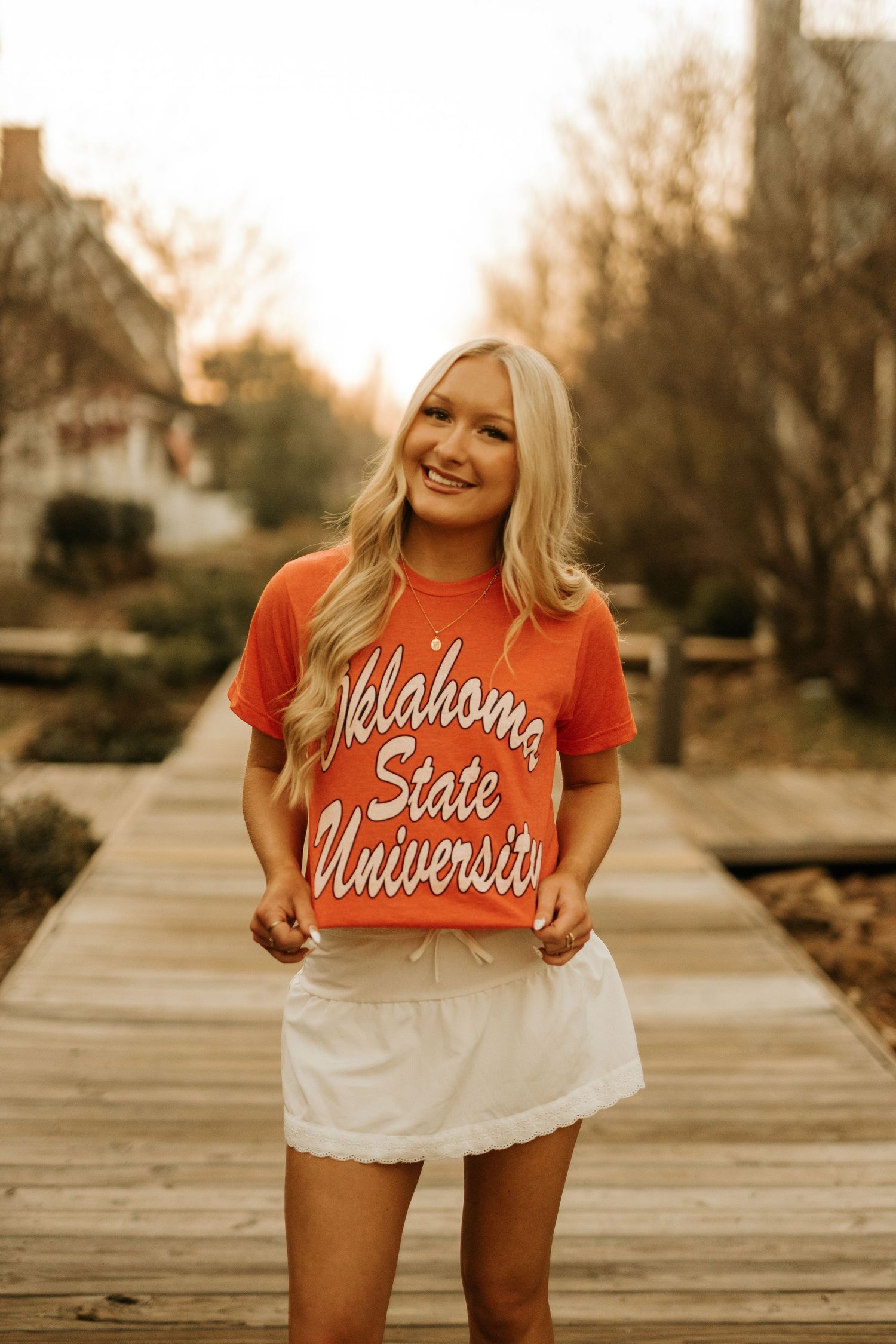 Smiling blonde woman wearing an orange Oklahoma State University t-shirt and white skirt on a wooden walkway.