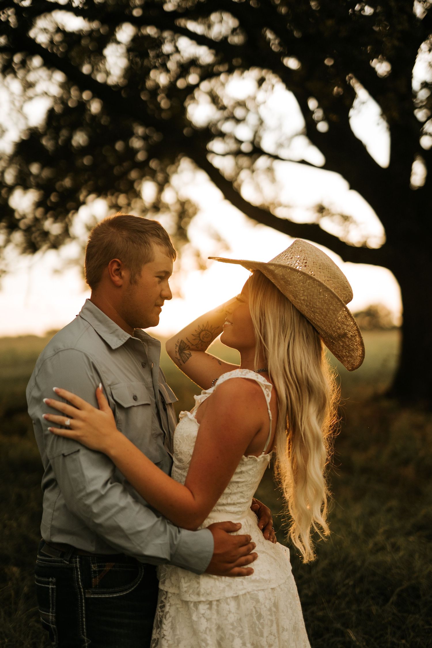 A couple shares a romantic sunset moment outdoors, woman in lace dress and cowboy hat, man in gray shirt embracing her.