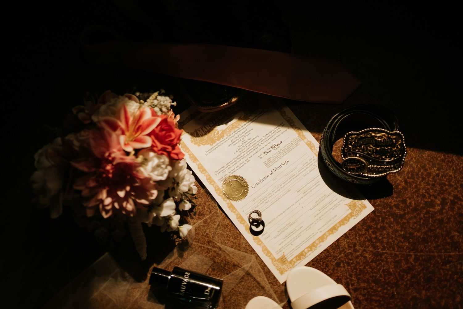 Wedding certificate with gold wax seal beside pink floral bouquet and rings on a dark surface.